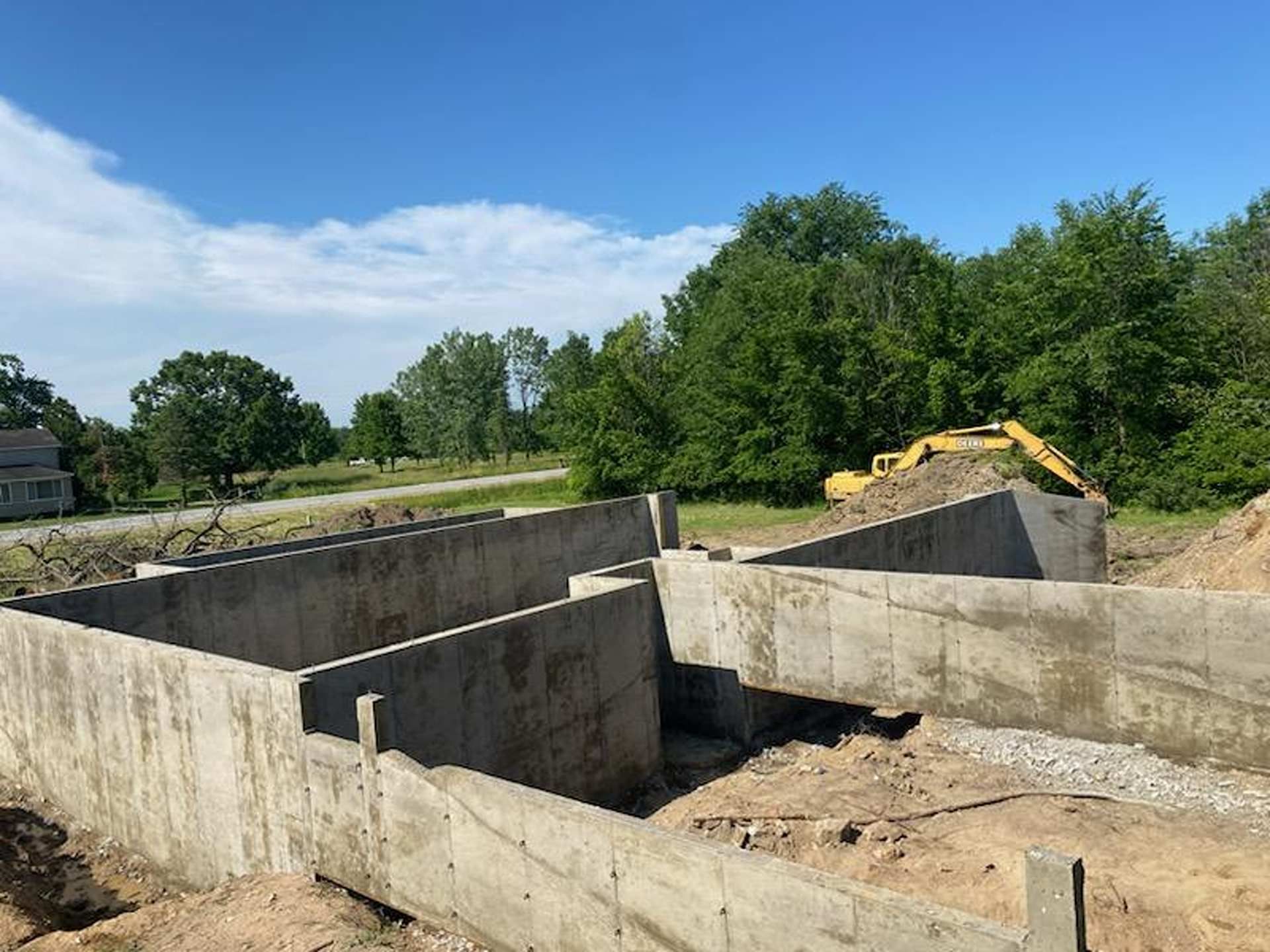 A concrete foundation for a building under construction on a sunny day with a yellow excavator in the background.