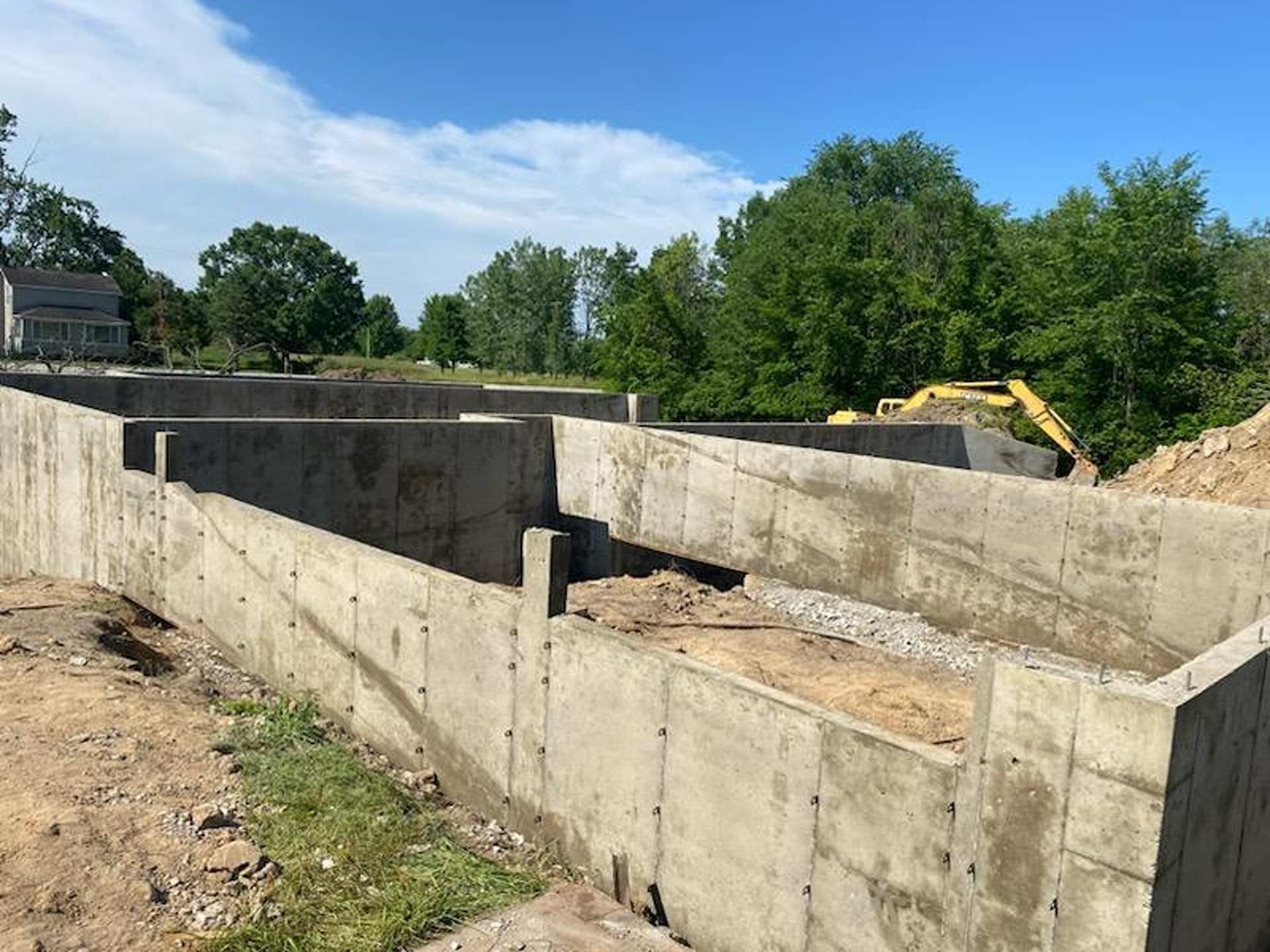 Concrete foundation walls for a new house under construction on a sunny day with trees and an excavator in the background.