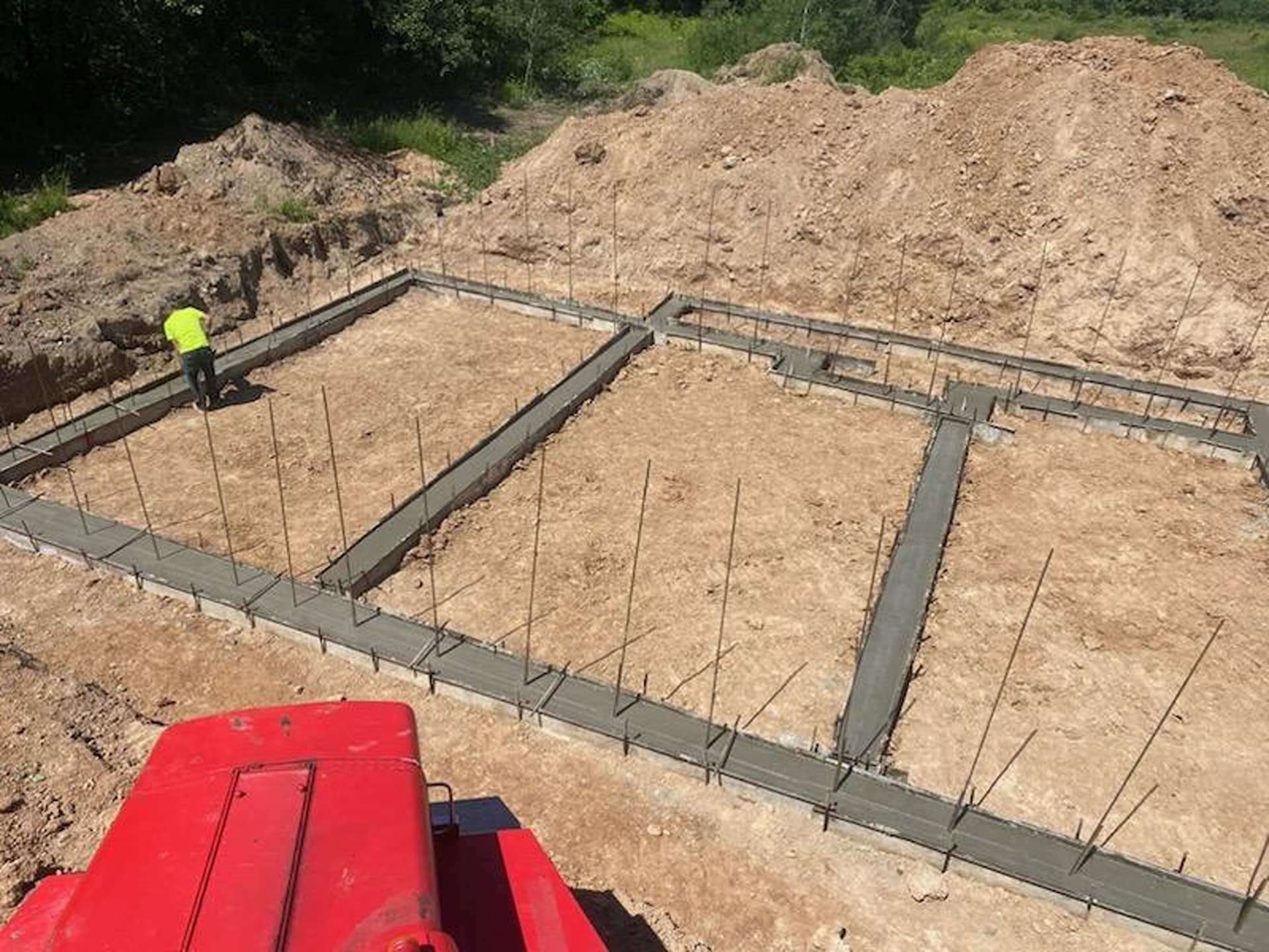 A person in a neon yellow shirt works on a concrete foundation trench at a construction site.