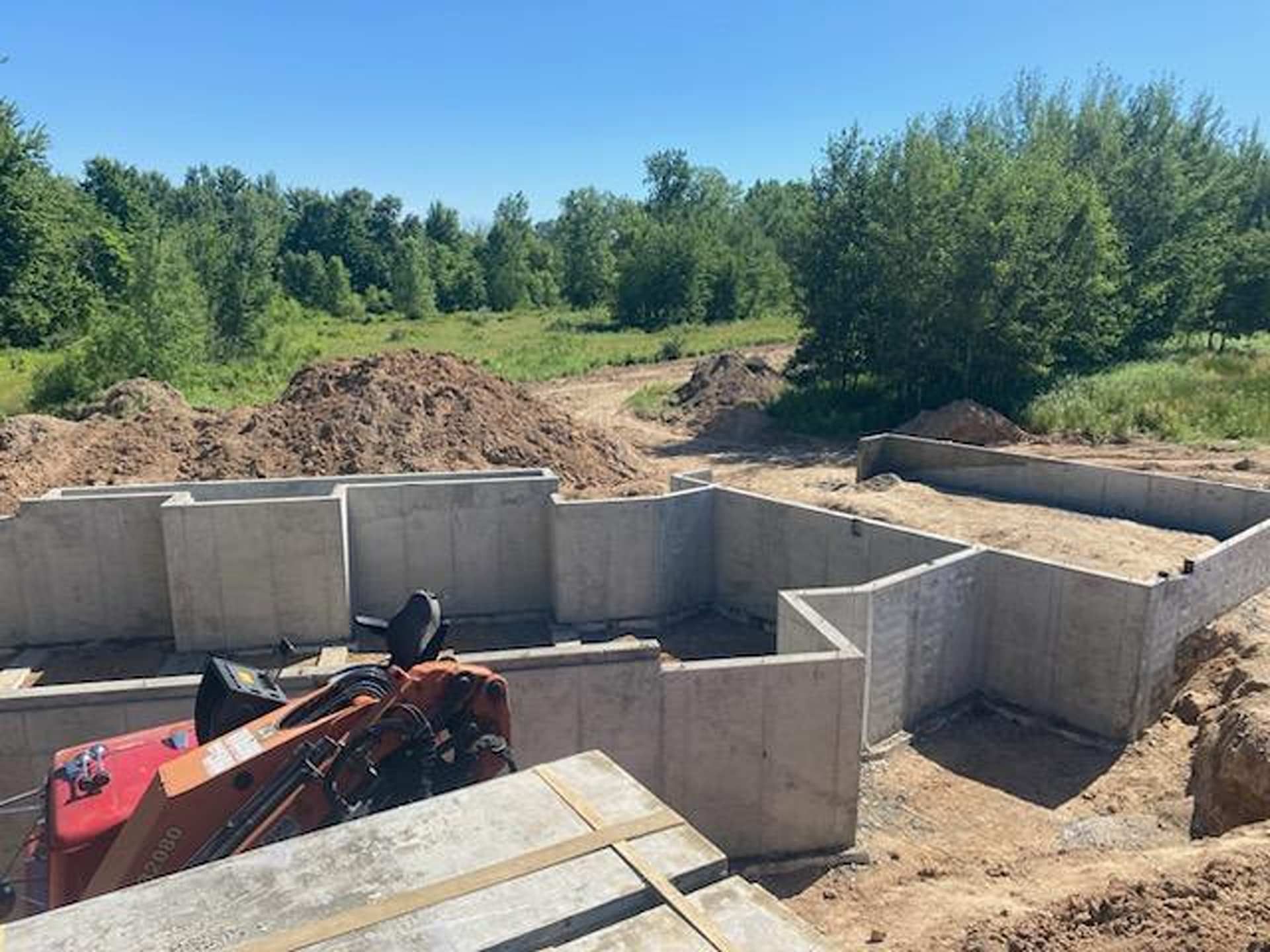 Concrete foundation walls for a new house under construction, with piles of dirt and equipment in a clearing.