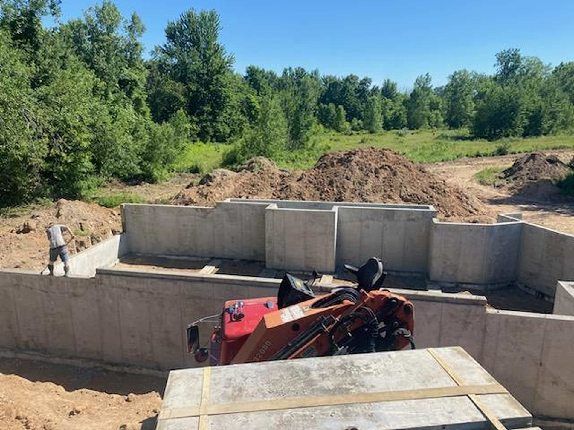 A worker stands in a concrete building foundation surrounded by dirt mounds and trees under a clear blue sky.