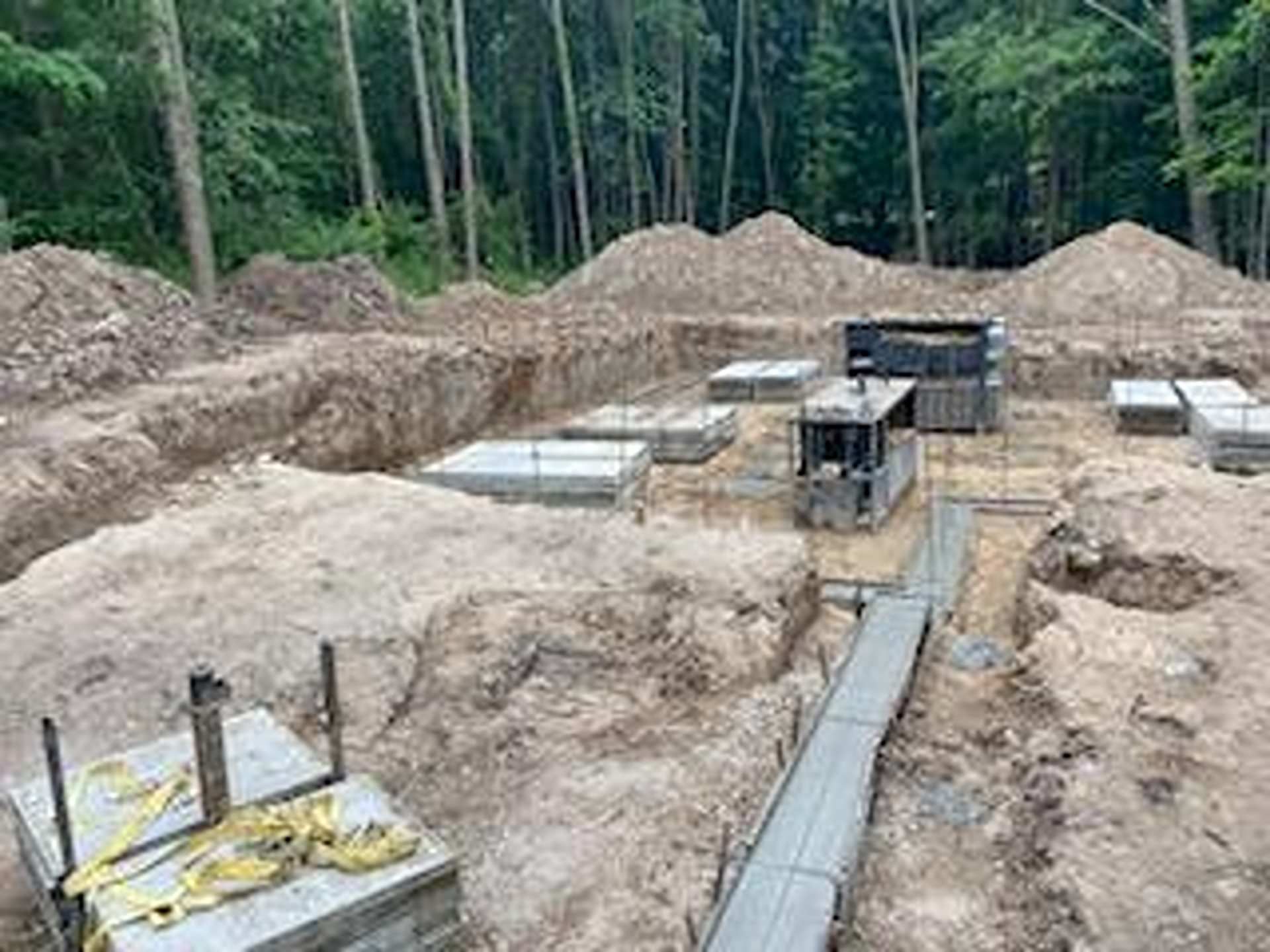 A construction site with excavated earth, concrete footings, and foundation forms set among trees.