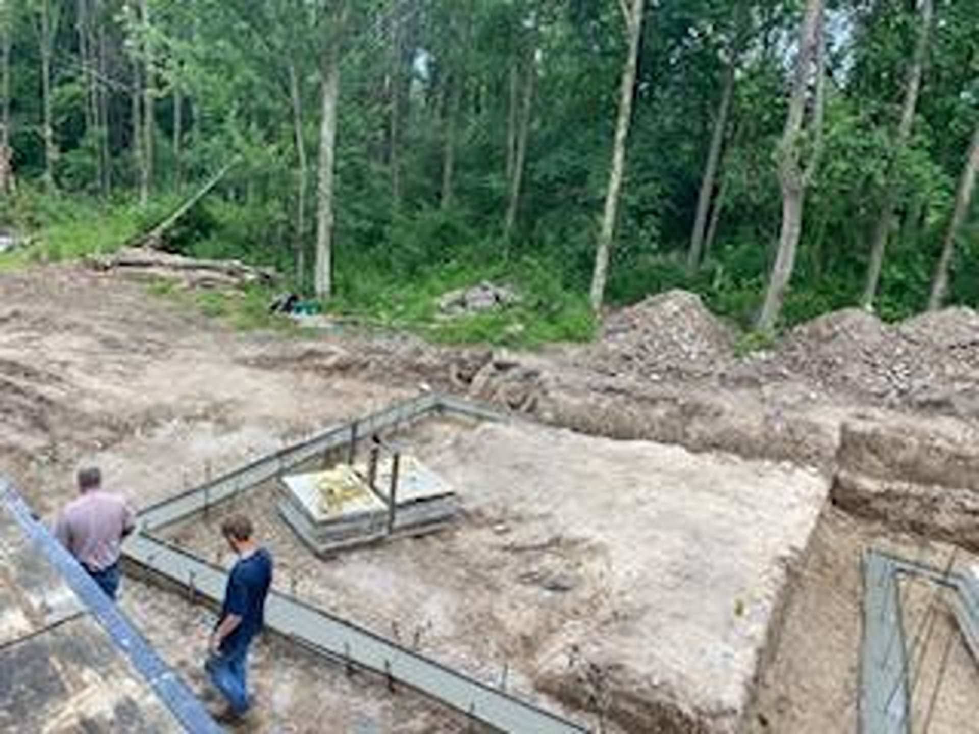Two people stand on a construction site overlooking a building foundation with metal rebar forms in a wooded area.