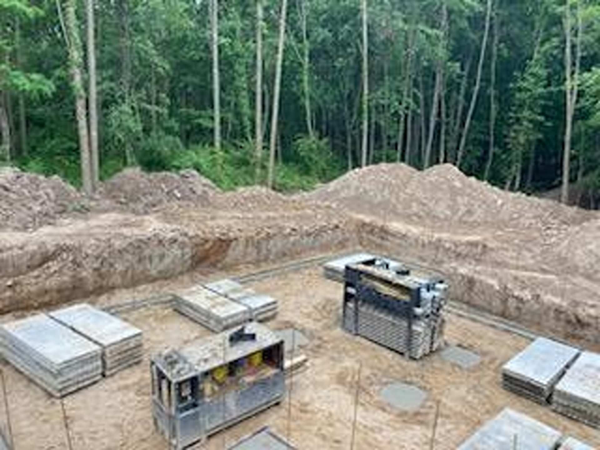 Construction site with piles of concrete blocks, metal machinery, and rebar footings set against a backdrop of woods.