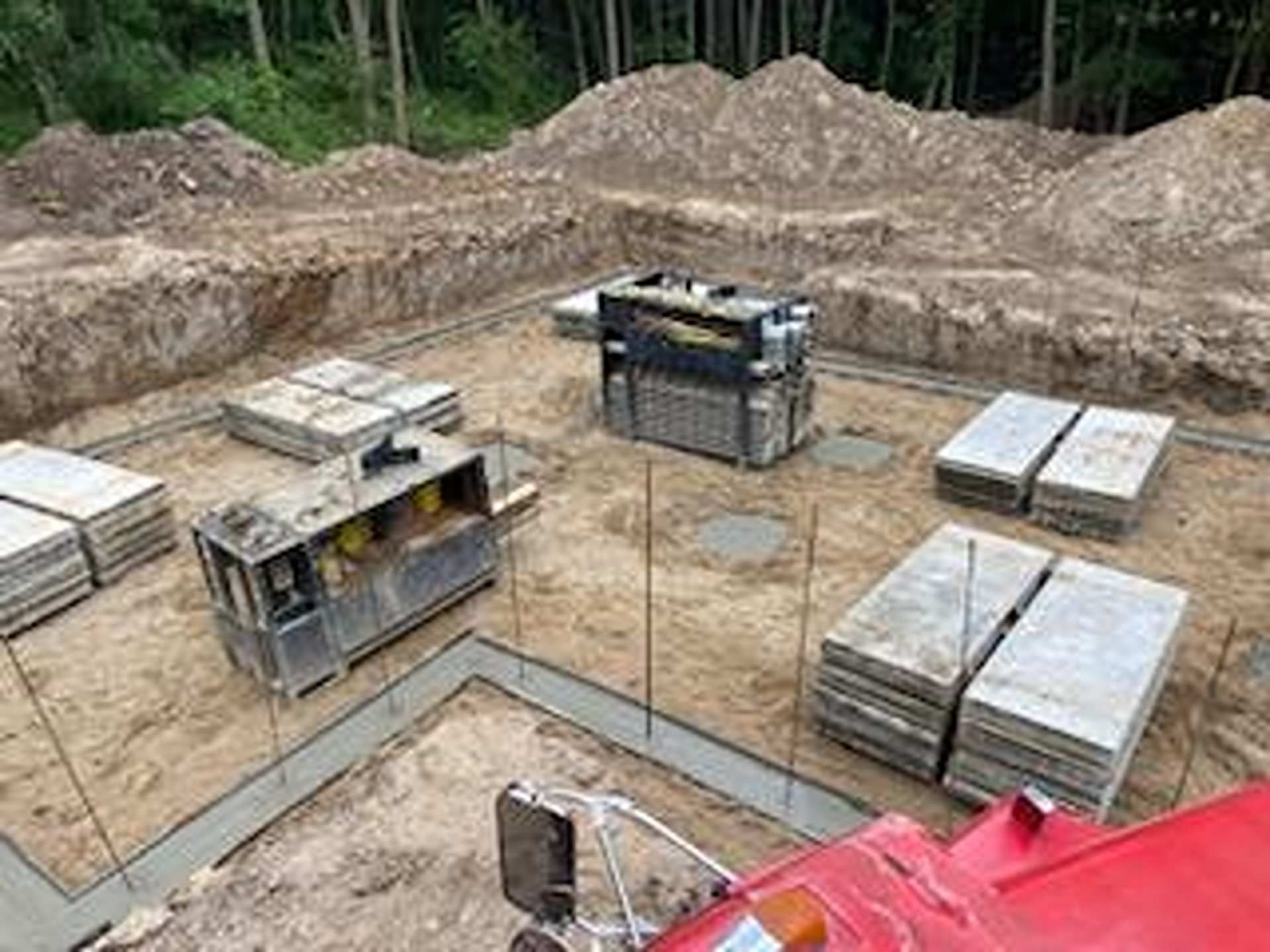 Construction site with concrete foundation walls, stacked paving stones, and metal equipment in a dirt excavation pit.