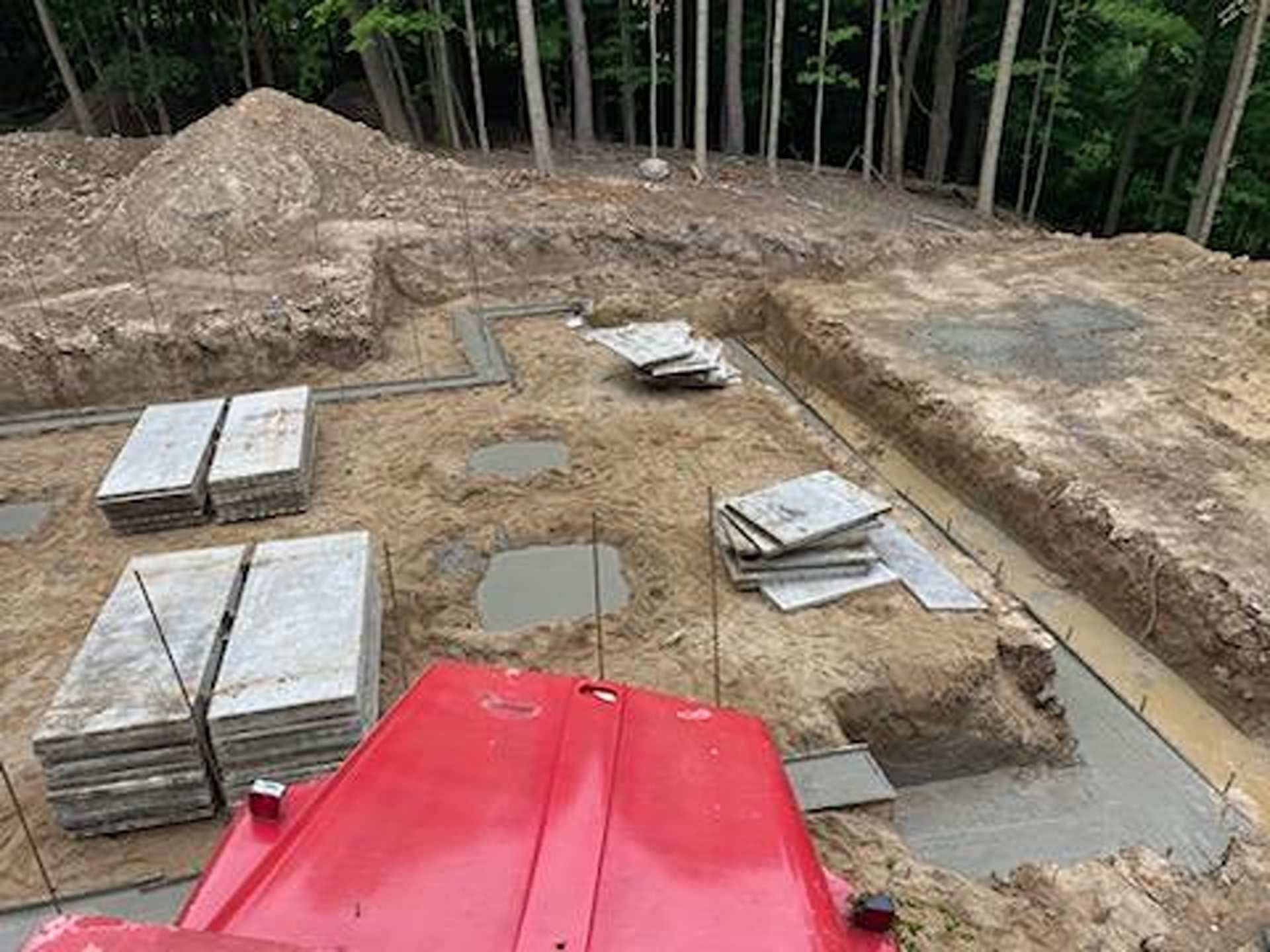 An aerial view of a construction site showing concrete foundations, stacks of gray pavers, and a red machine in the foreground.