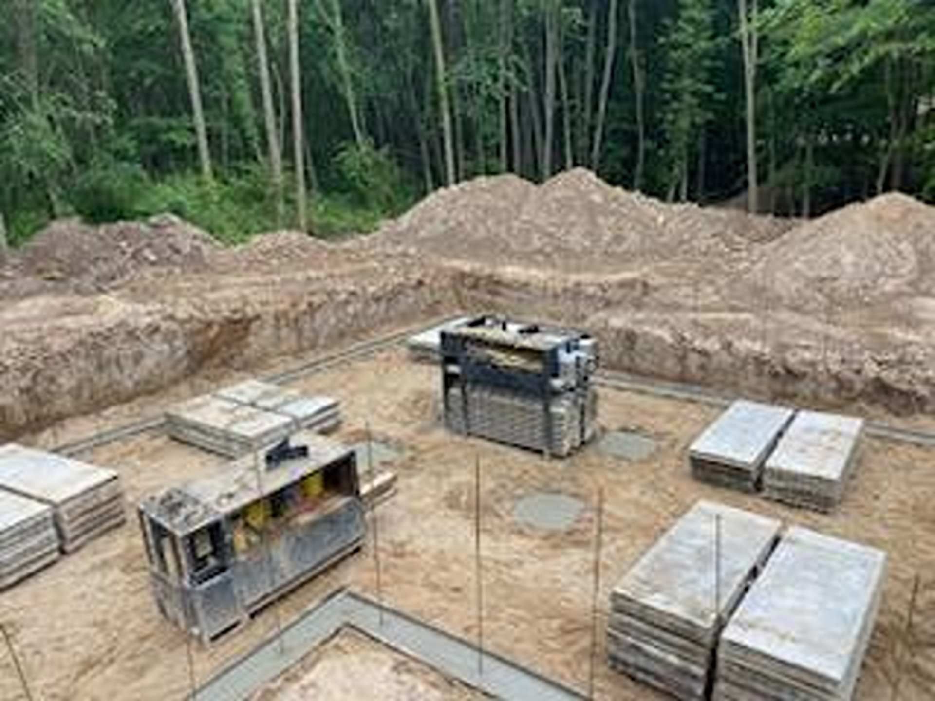 A construction site showing a concrete foundation trench with stacks of stone pavers and building equipment on sandy soil.