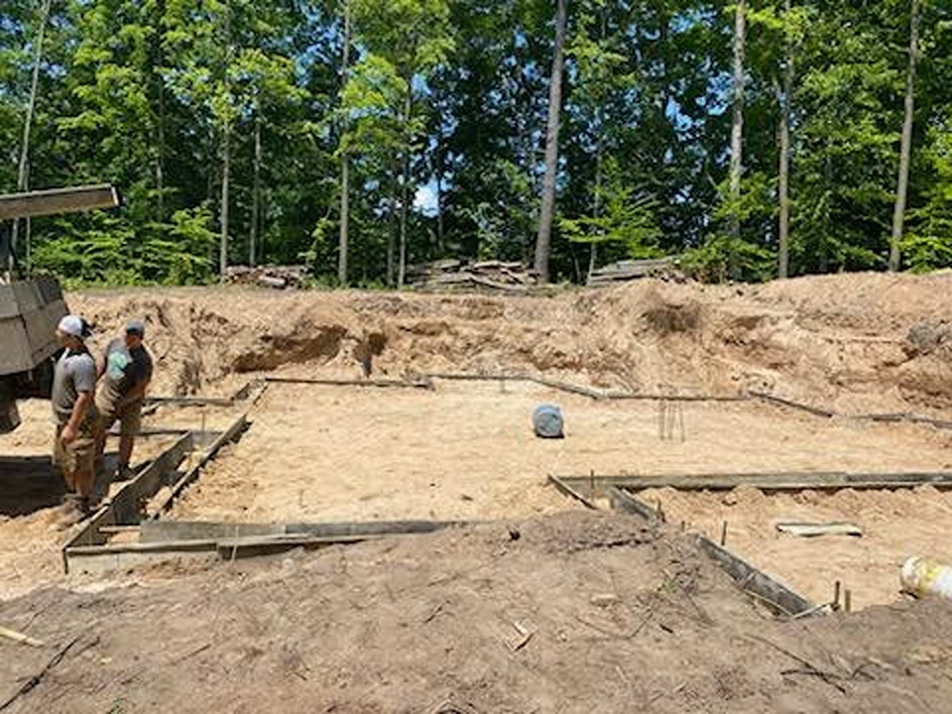 Two people stand at a construction site with wooden concrete forms laid out in a large, excavated sandy area.