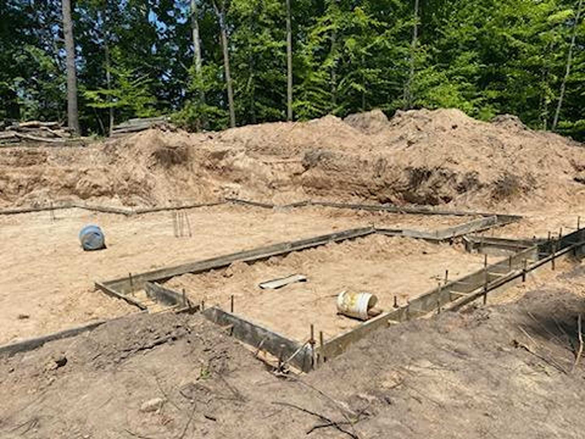 Wooden foundation forms for a new building under construction, surrounded by sandy soil and a forest backdrop.