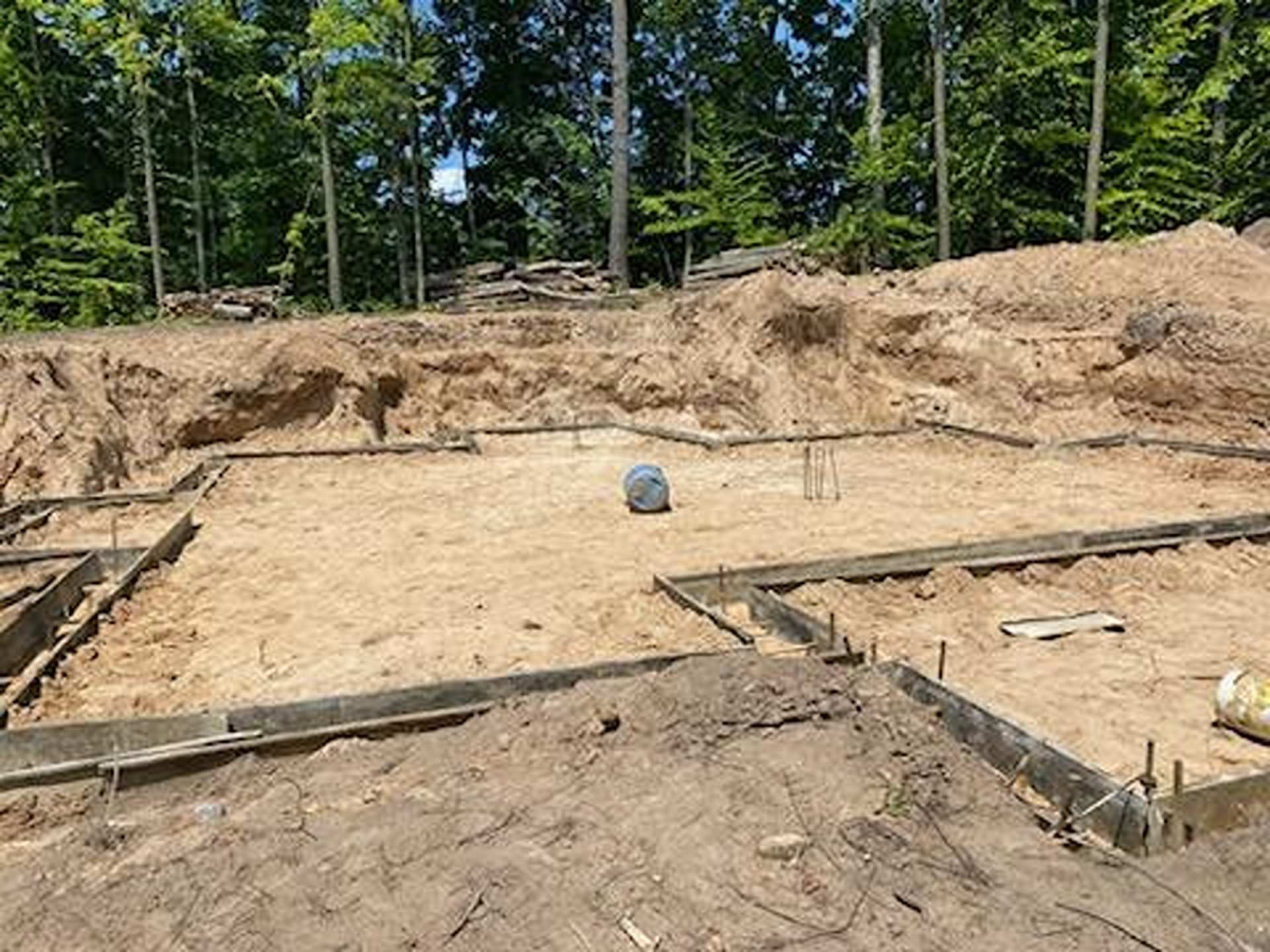 A construction site showing wooden foundation forms set in a sandy excavated lot with a forest in the background.