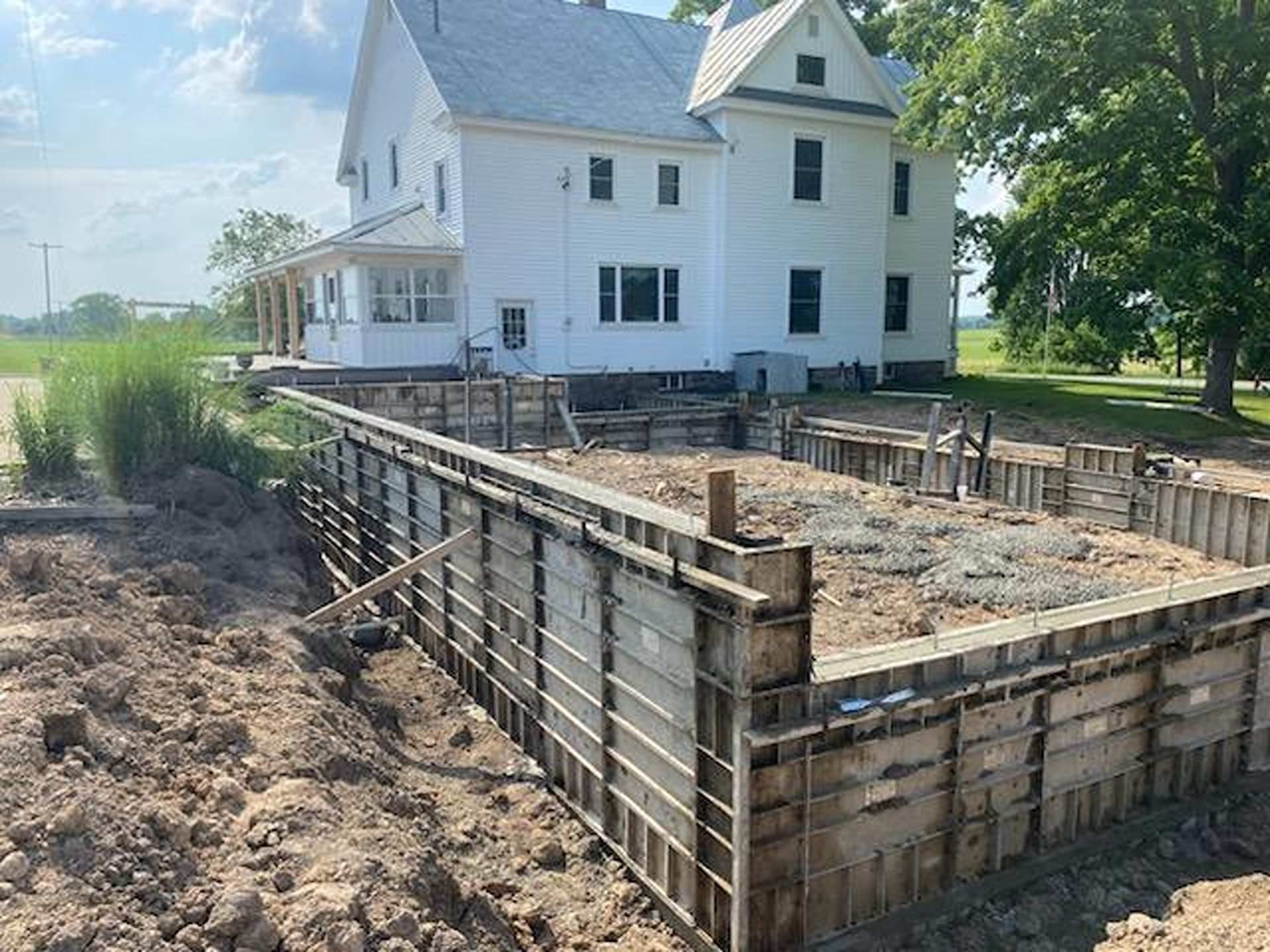 A two-story white farmhouse stands behind a large concrete foundation under construction with metal framing forms.