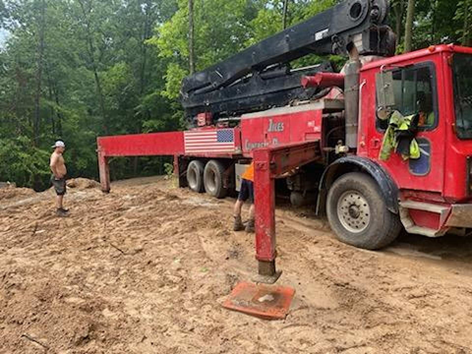 A red crane truck with its outriggers extended is parked on a dirt lot near a wooded area, with a person standing nearby.