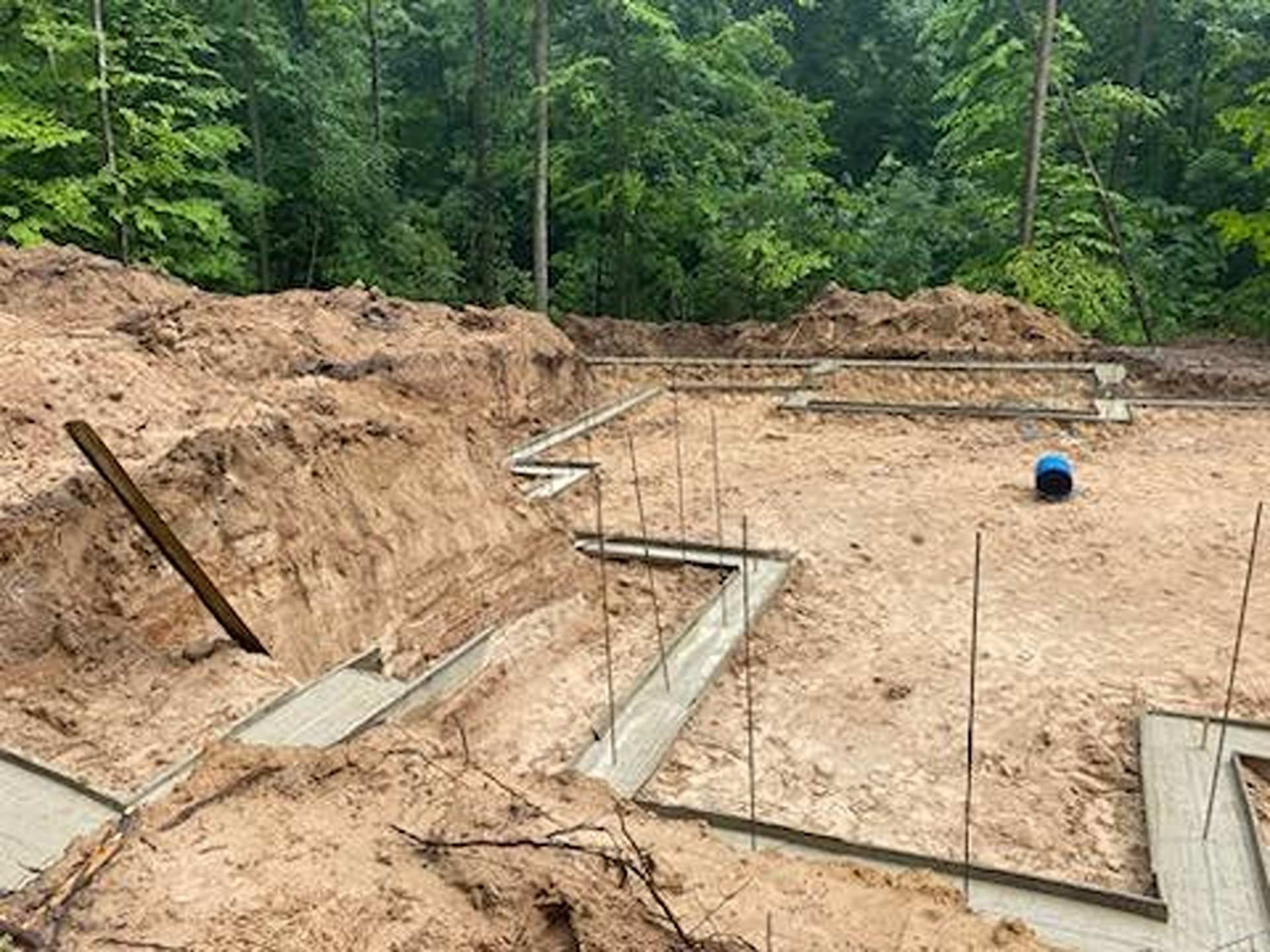 Concrete foundation footings for a building under construction, surrounded by piles of dirt and a forested background.