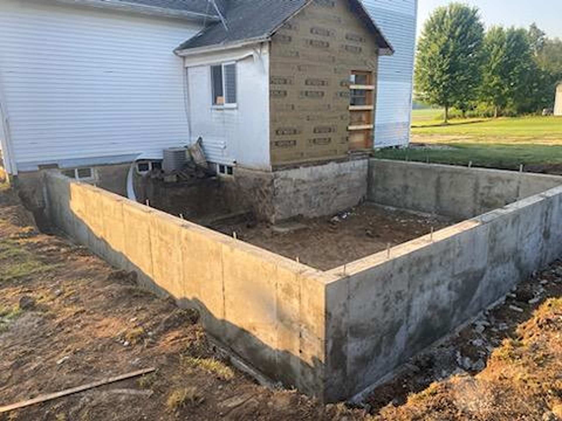A concrete foundation wall newly constructed for a home addition, positioned against the side of an existing white house.