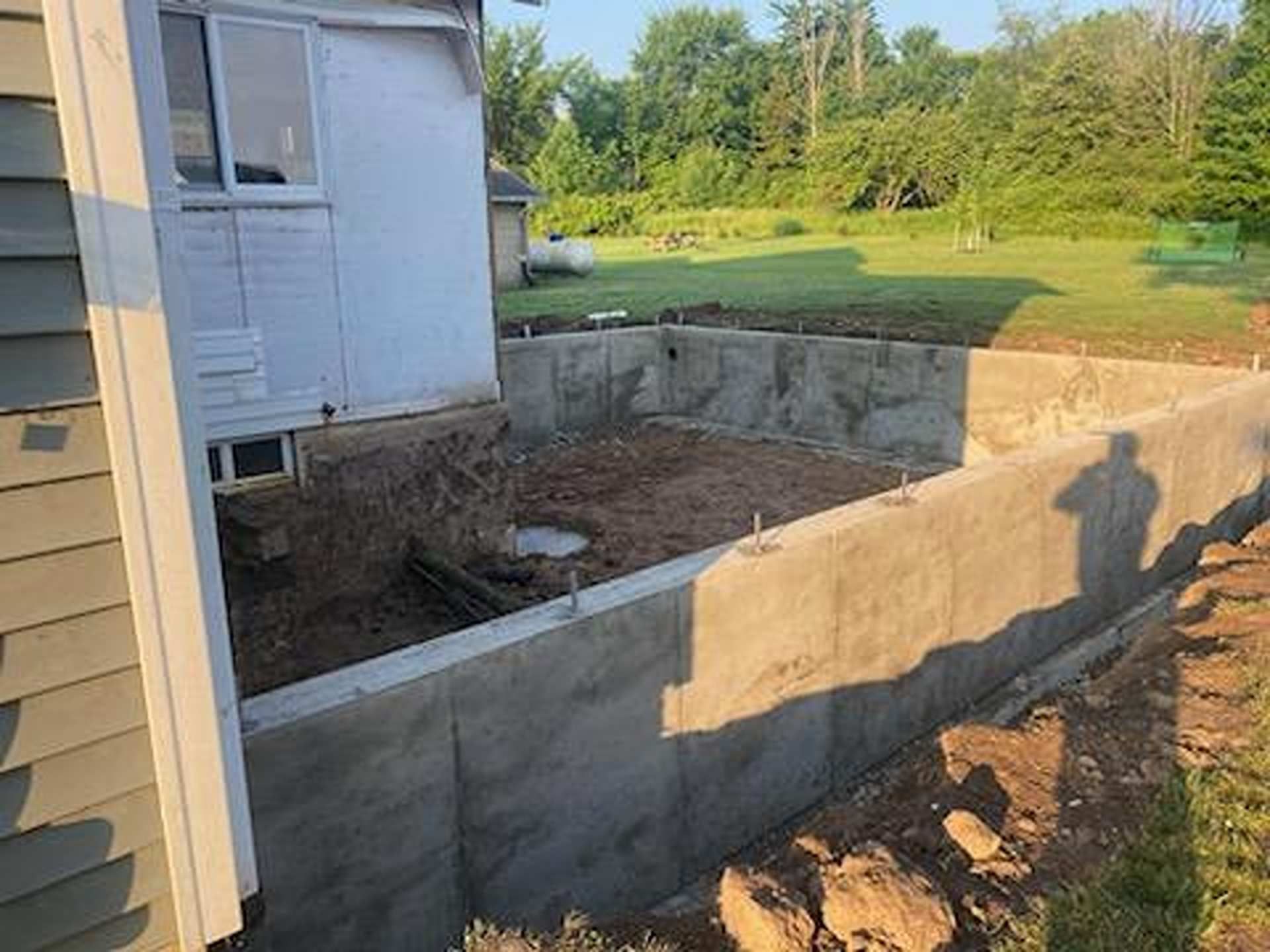 A concrete foundation wall for a new home addition stands next to the side of a house on a sunny, grassy lot.
