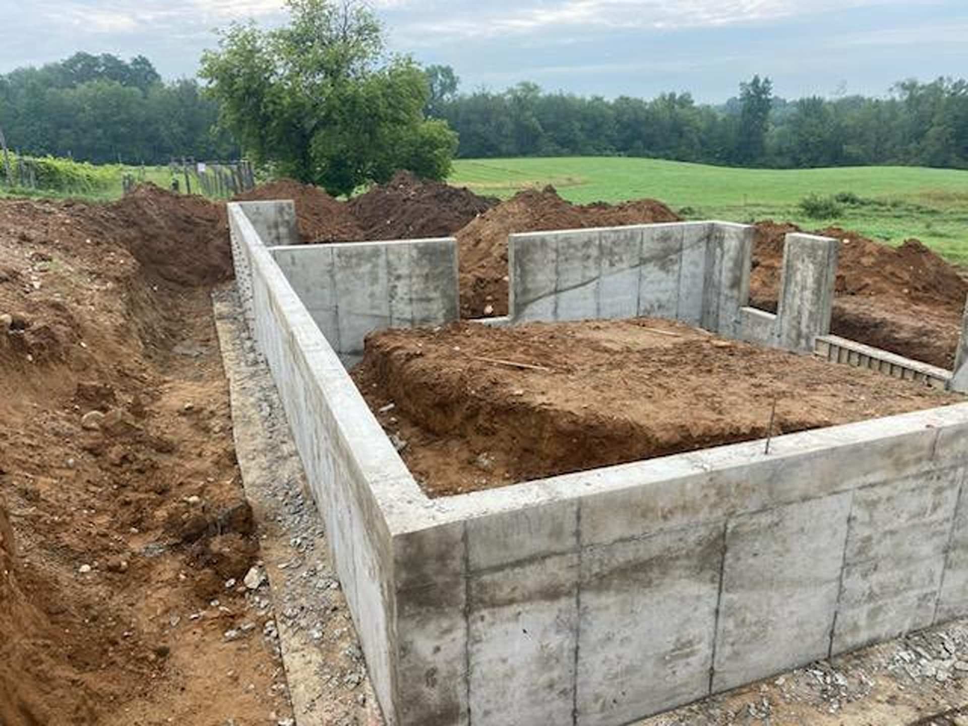 Concrete foundation walls of a building under construction, surrounded by piles of dirt in a grassy field.