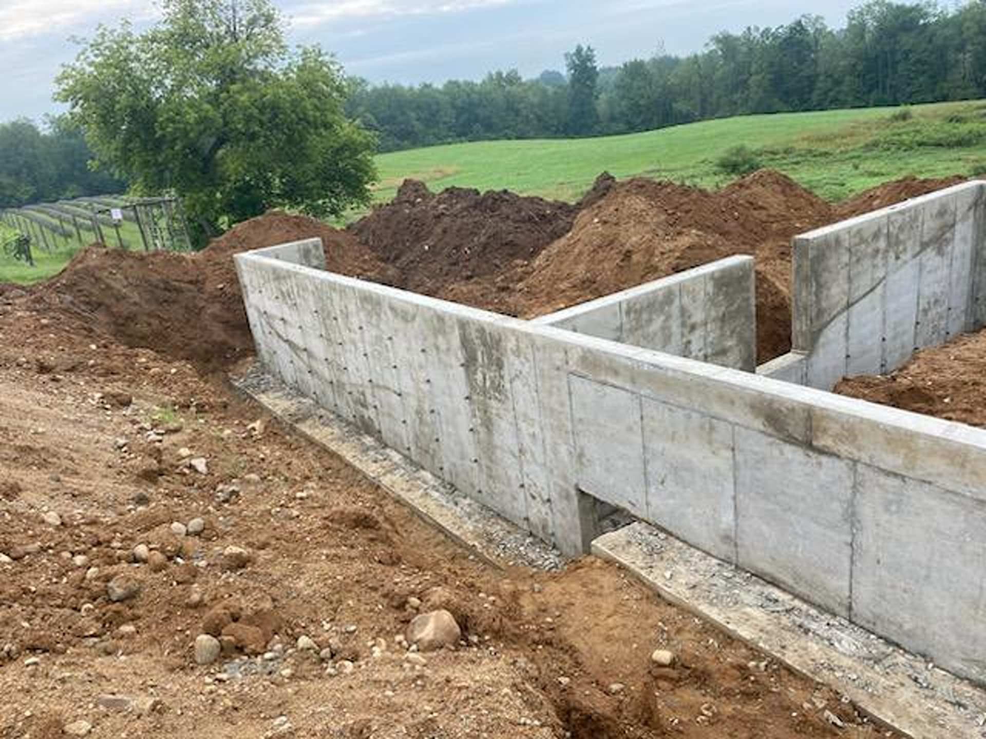 Concrete foundation walls for a building under construction, surrounded by piles of dirt in a grassy field.