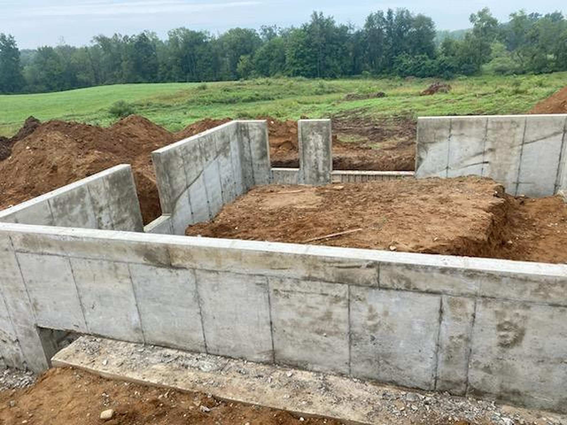 Concrete foundation walls for a new building under construction in a grassy field with piles of excavated dirt.