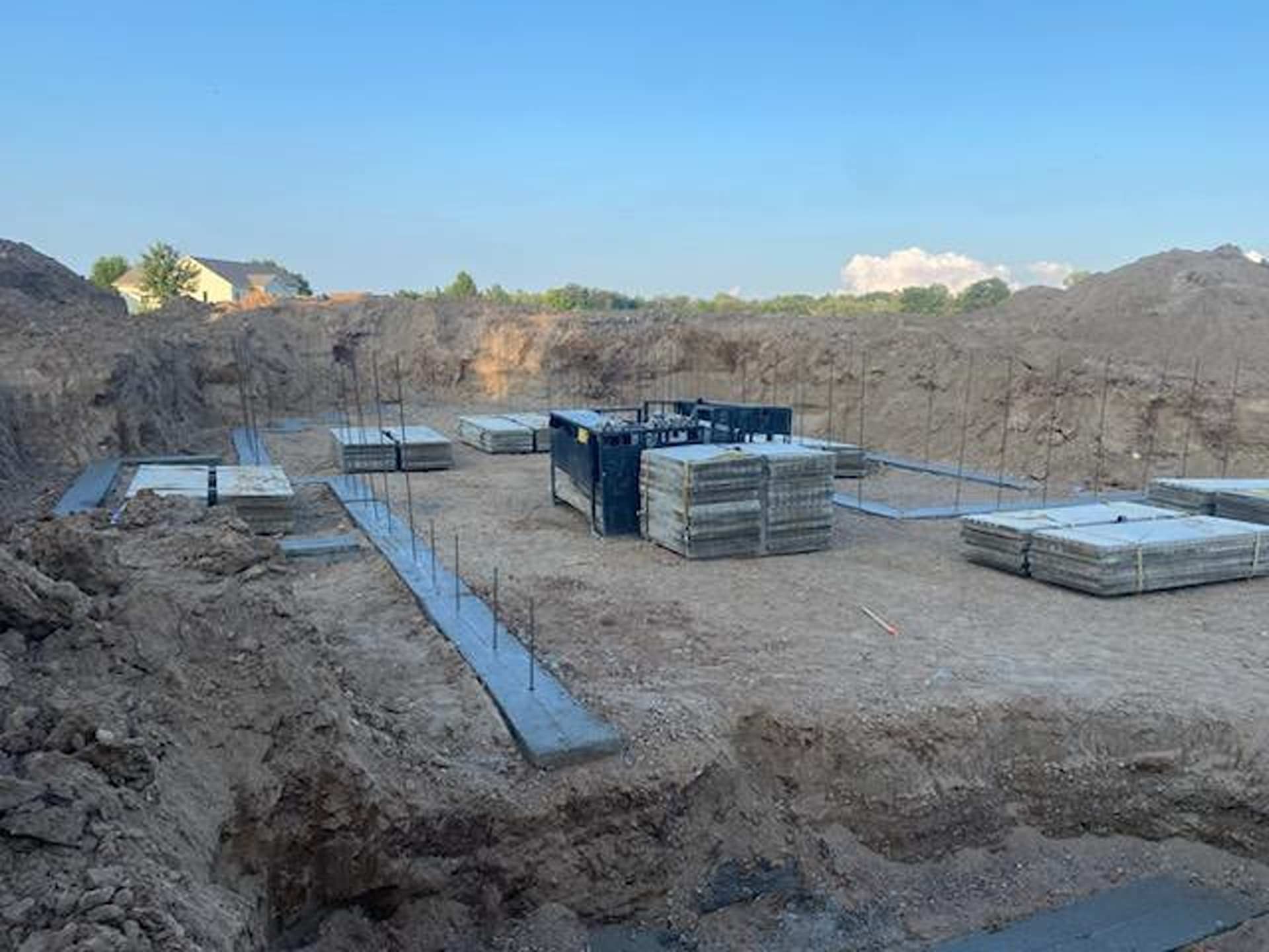 A construction site showing concrete foundation forms and stacks of paving stones in a dirt excavation pit.