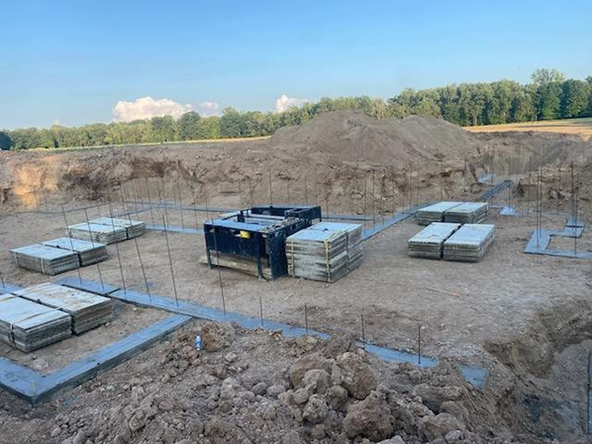 A construction site with poured concrete footings, stacks of concrete blocks, and vertical rebar in a dirt excavation.