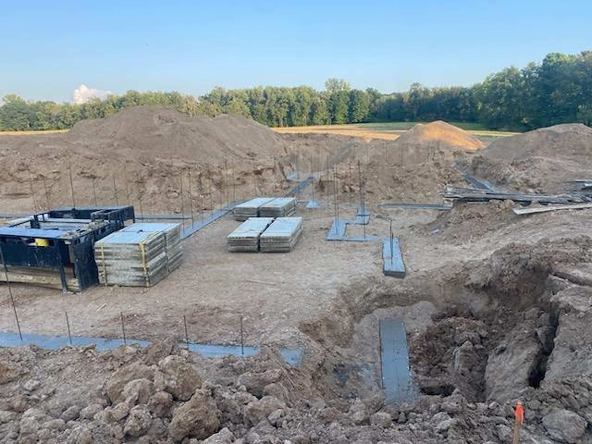 A construction site with excavated dirt, concrete footings, and stacks of construction materials under a clear blue sky.