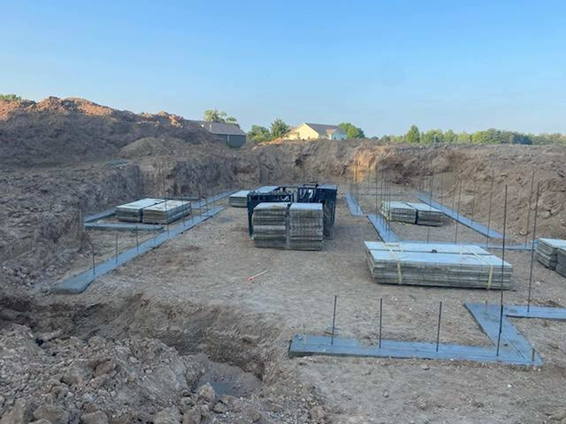 A construction site shows concrete foundation footings in a large excavated dirt pit, with stacks of paving stones.