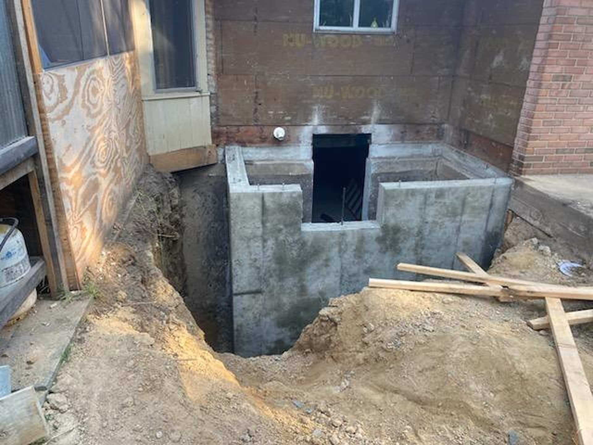 A newly poured concrete foundation structure sits in an excavated dirt pit next to a brick building.