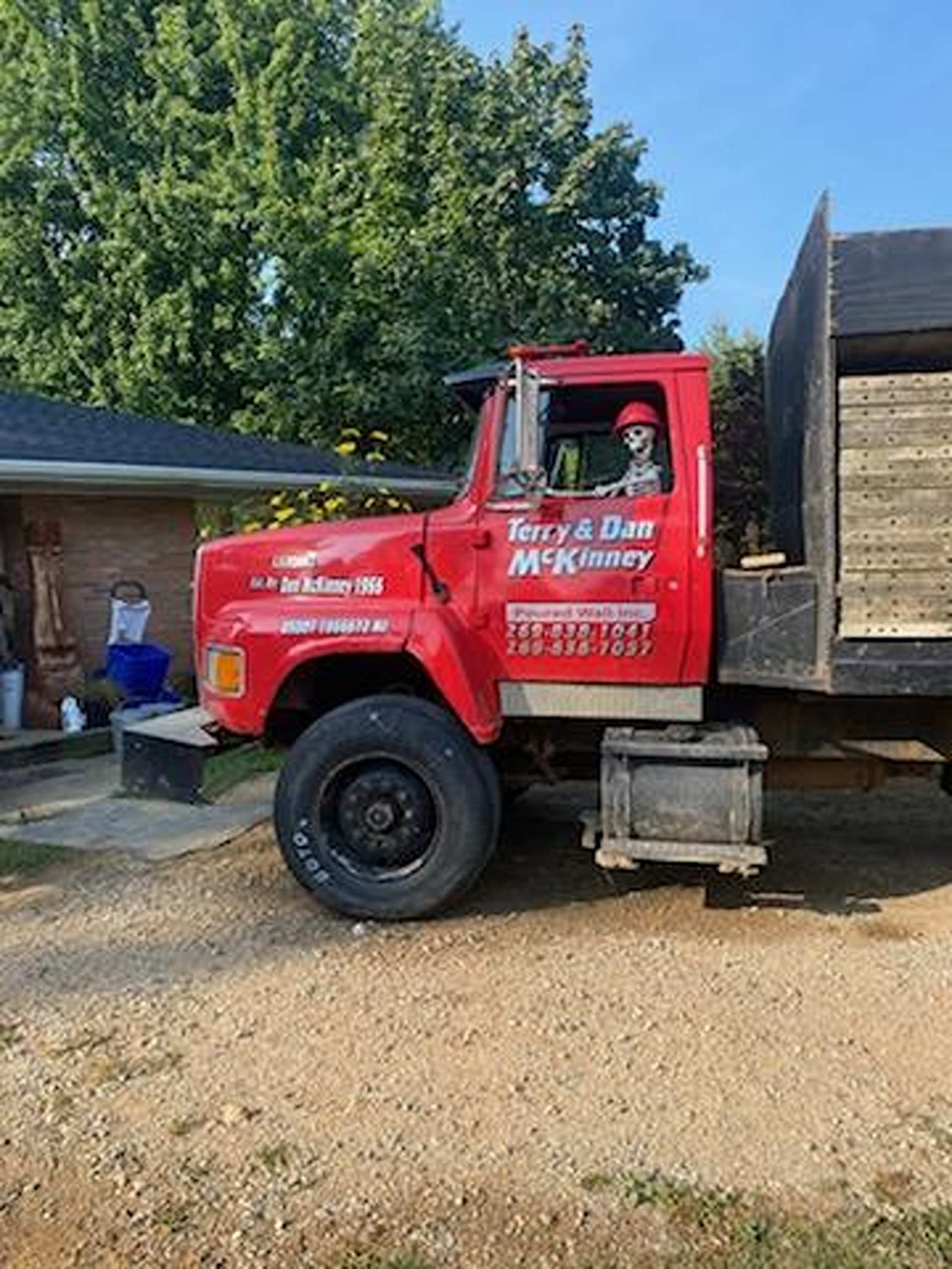 A red dump truck parked on a gravel driveway, with a decorative skeleton wearing a hard hat sitting in the driver's seat.