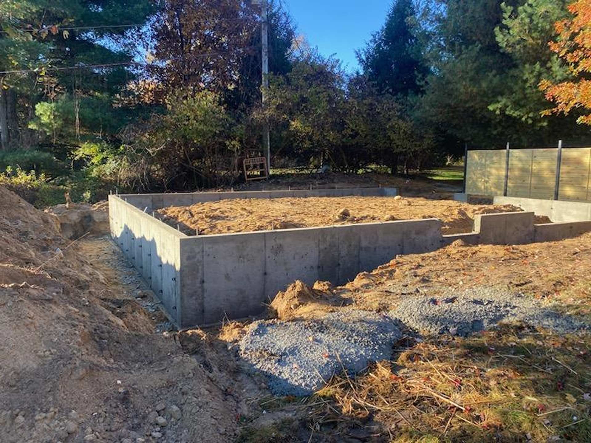A concrete foundation for a building under construction, surrounded by piles of dirt and gravel in a wooded area.