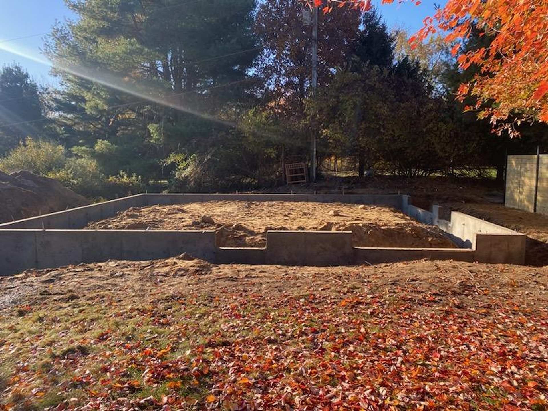 Concrete foundation walls for a building under construction on a dirt lot covered with fall leaves and surrounded by trees.