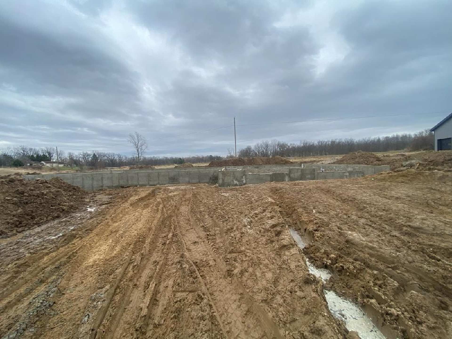 A wide, muddy construction site under a cloudy sky, featuring concrete foundation walls and deep tire tracks in the earth.