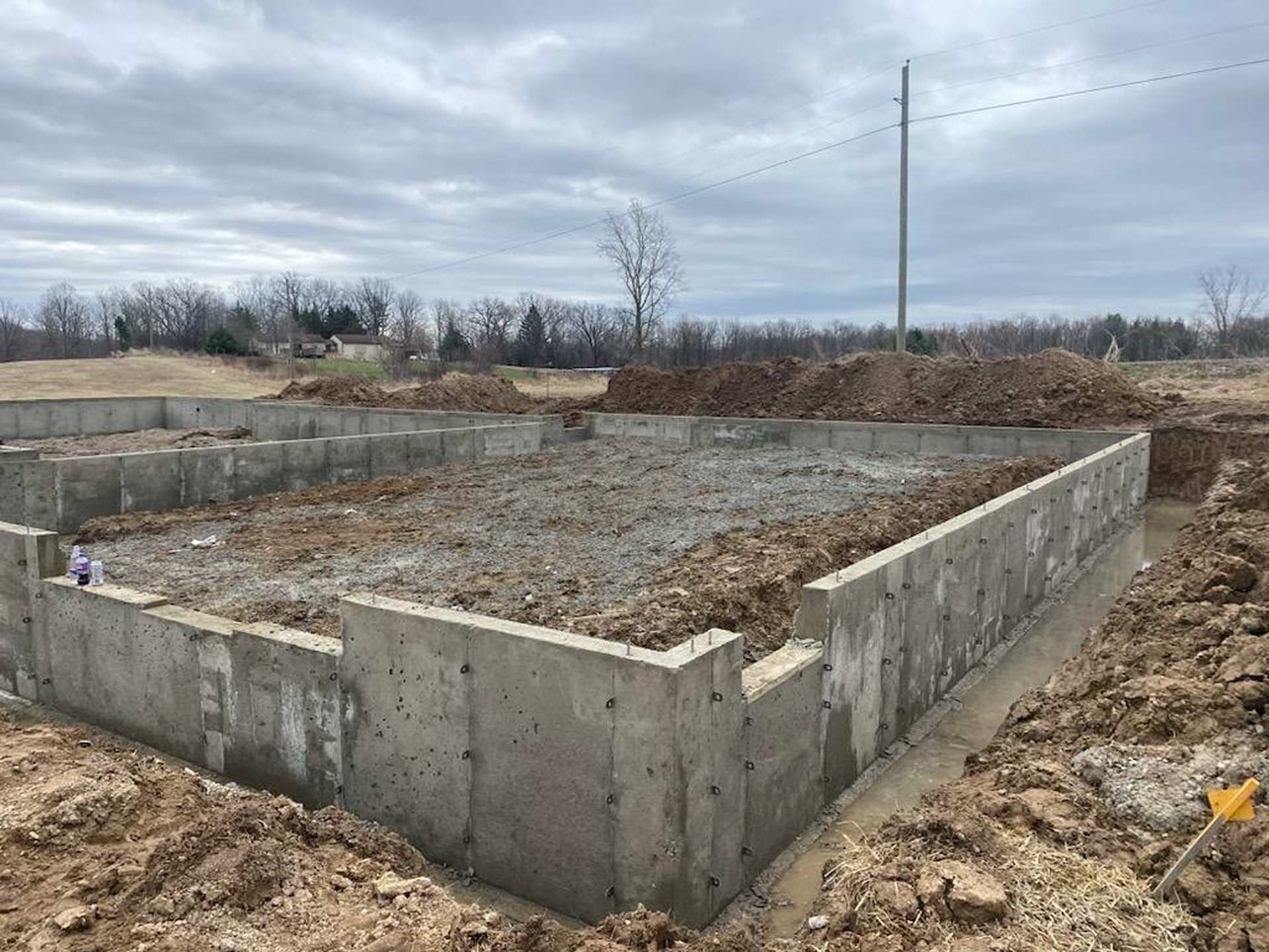 A poured concrete foundation for a new house construction, surrounded by piles of excavated dirt under a cloudy sky.