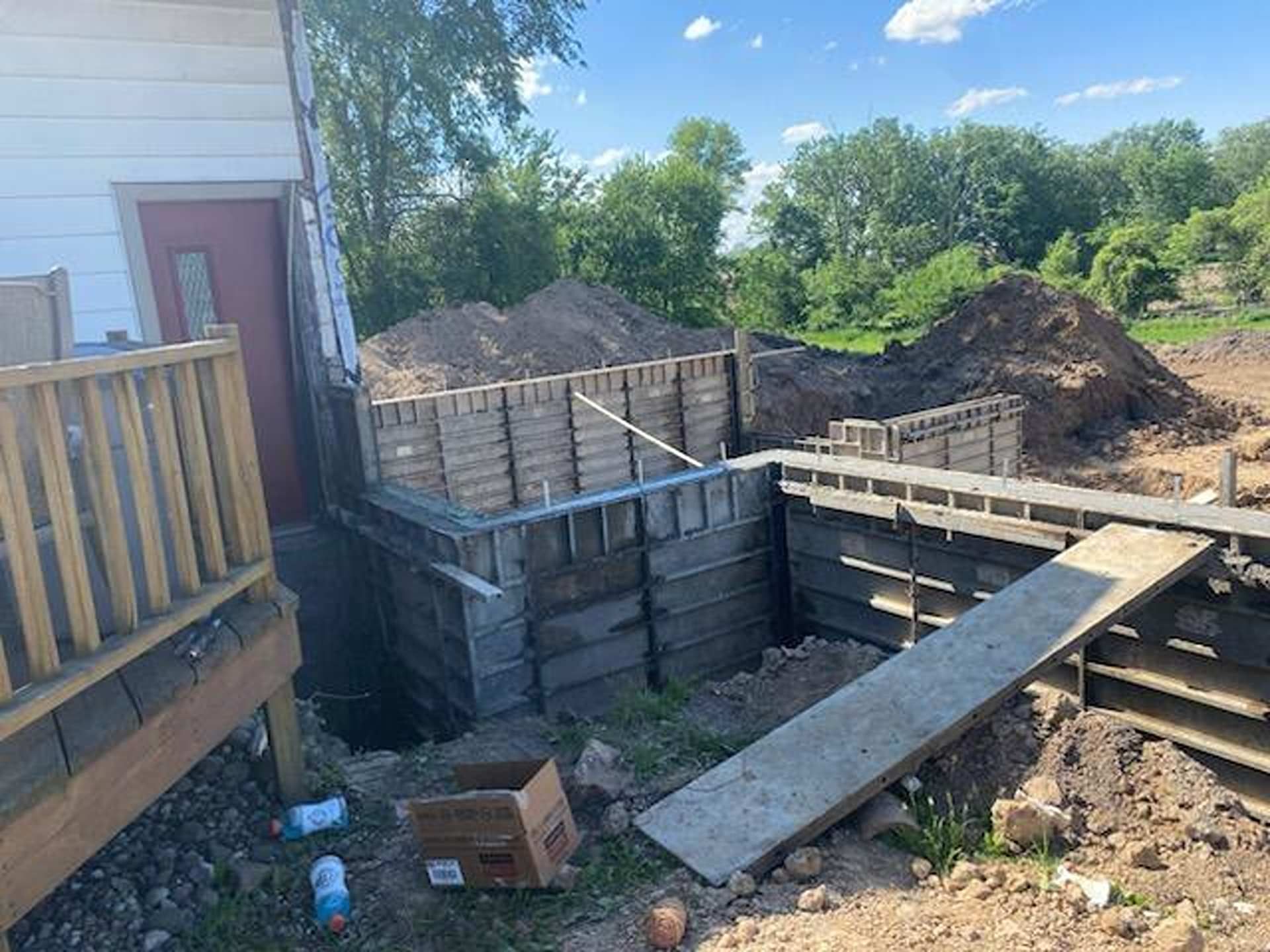 Construction site with concrete foundation forms next to a house with a wooden deck and piles of dirt in the background.