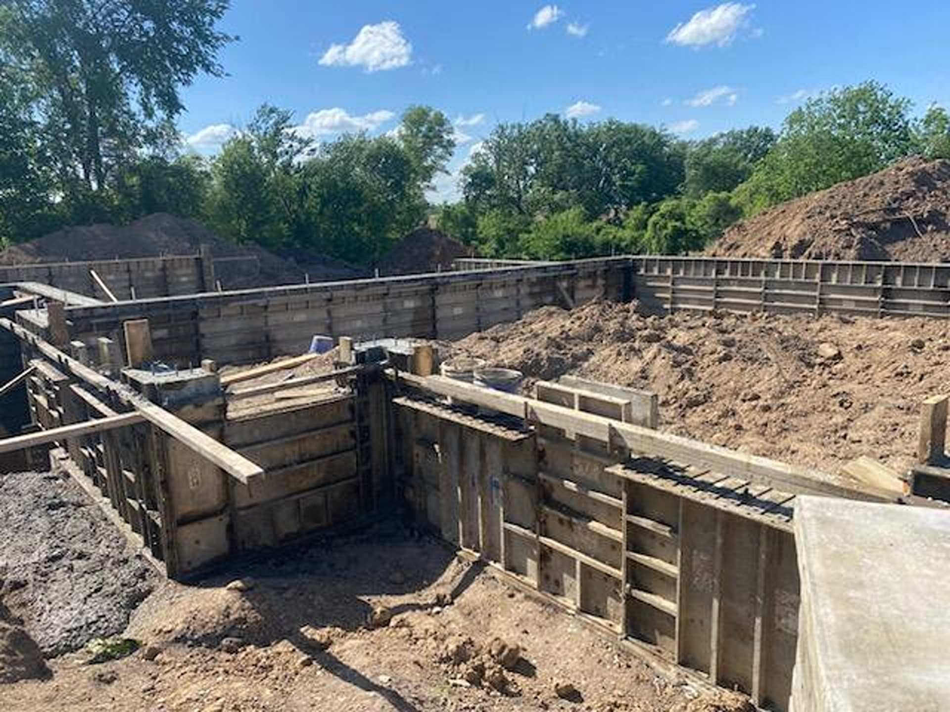 Wooden concrete forms set up for a house foundation at a sunny construction site with dirt piles and trees.