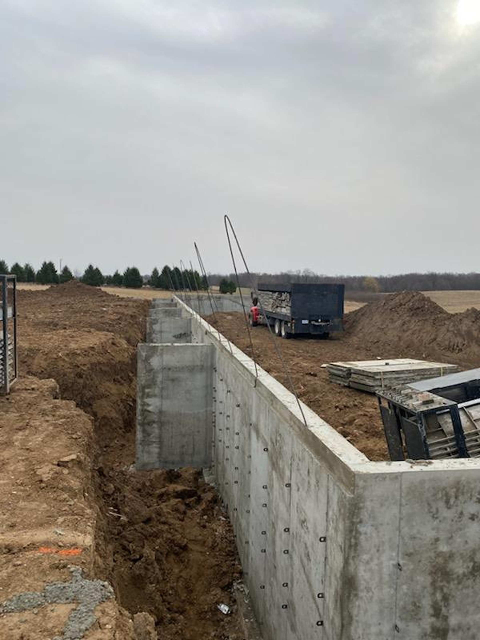 Concrete foundation walls in an open excavation site, with a truck parked in the background under a cloudy sky.