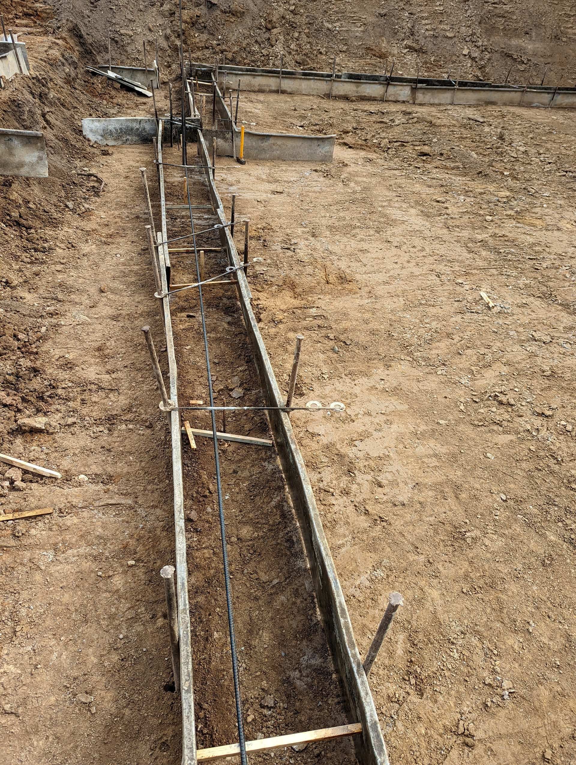 A trench for a building foundation with metal rebar forms and wooden stakes set in dirt at a construction site.
