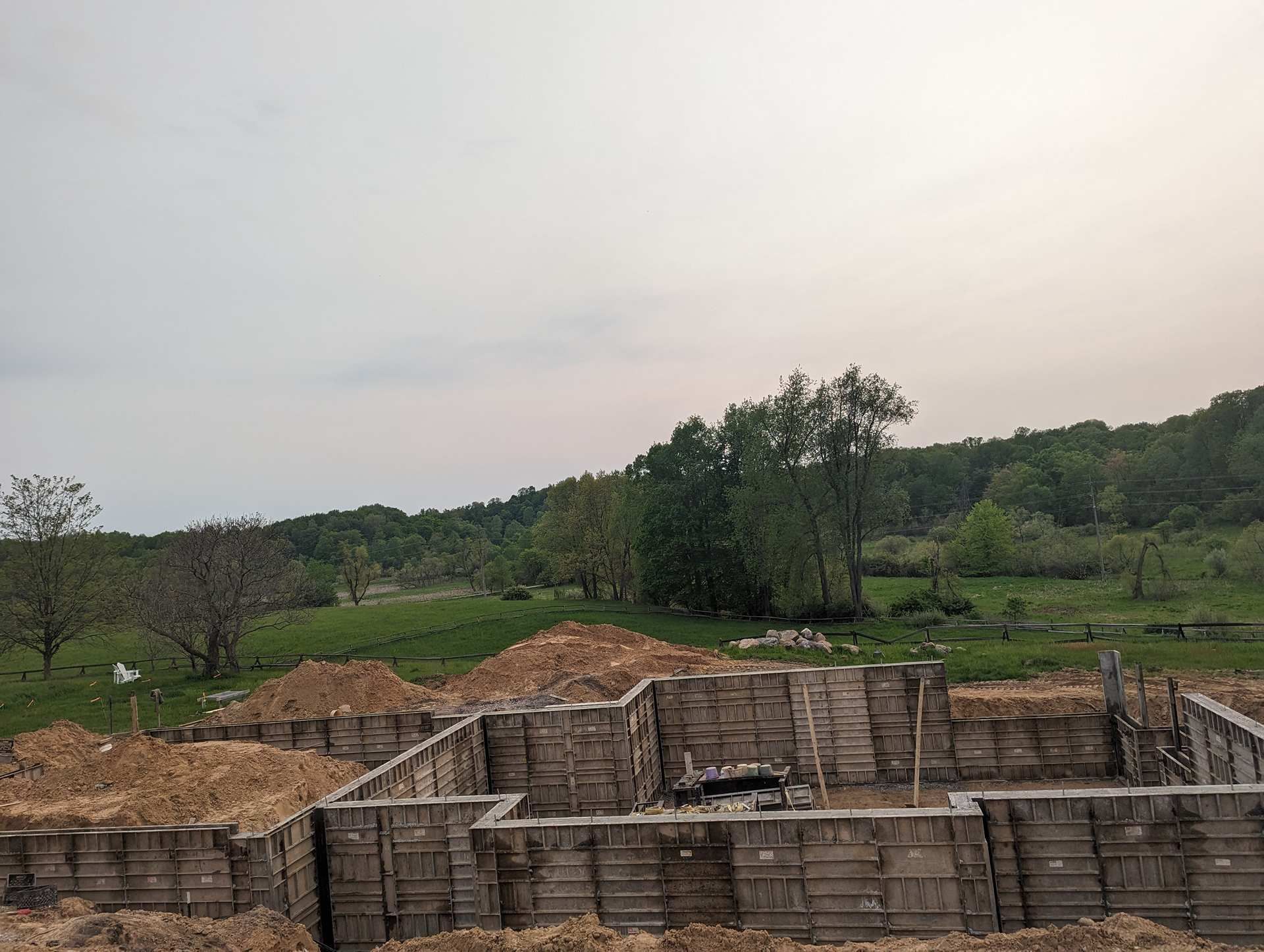 Wooden concrete formwork for a foundation on a dirt lot, set against a background of green trees and a cloudy sky.