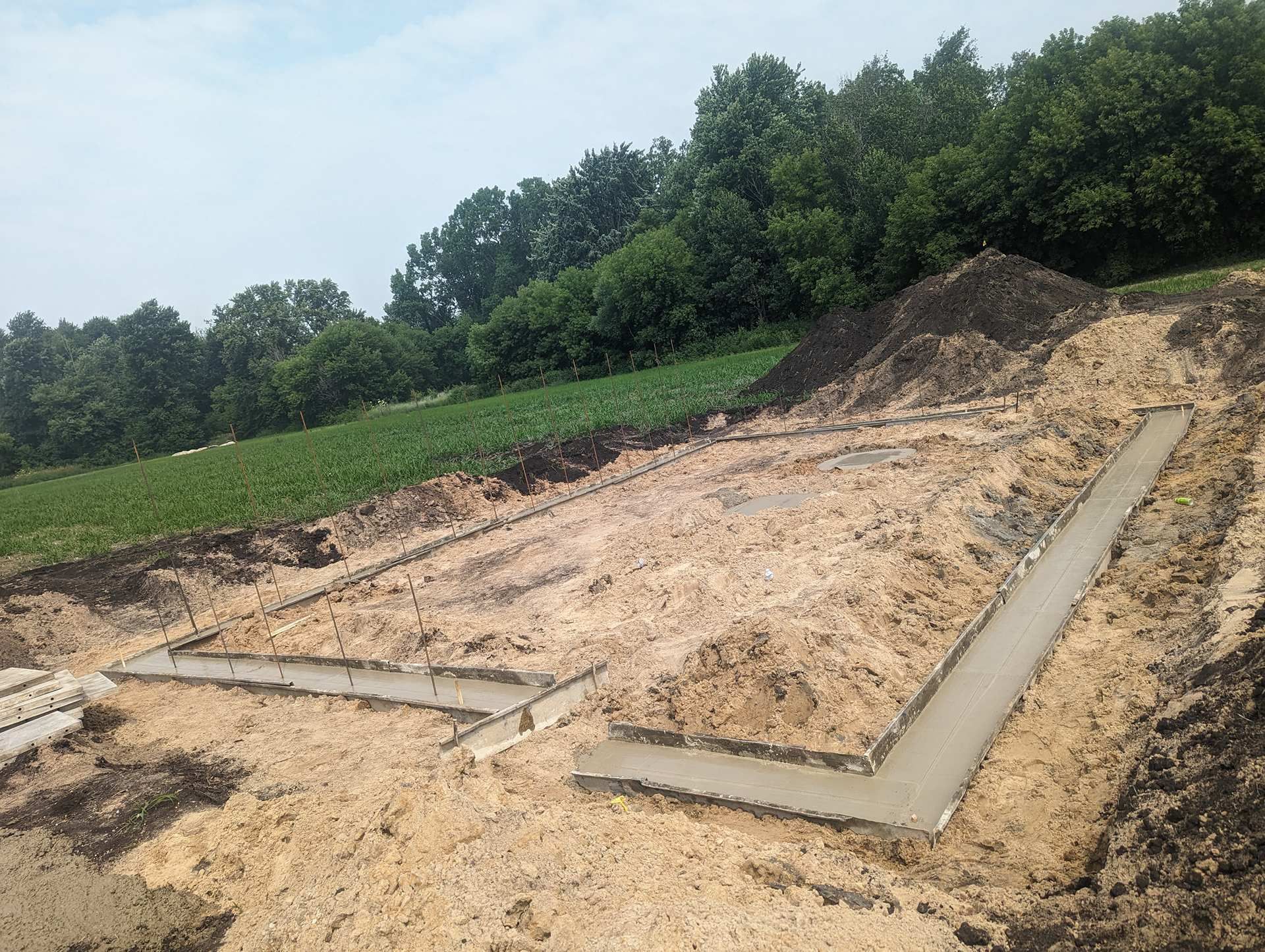Concrete footings for a building foundation set in a dirt trench, surrounded by a field and trees under a blue sky.