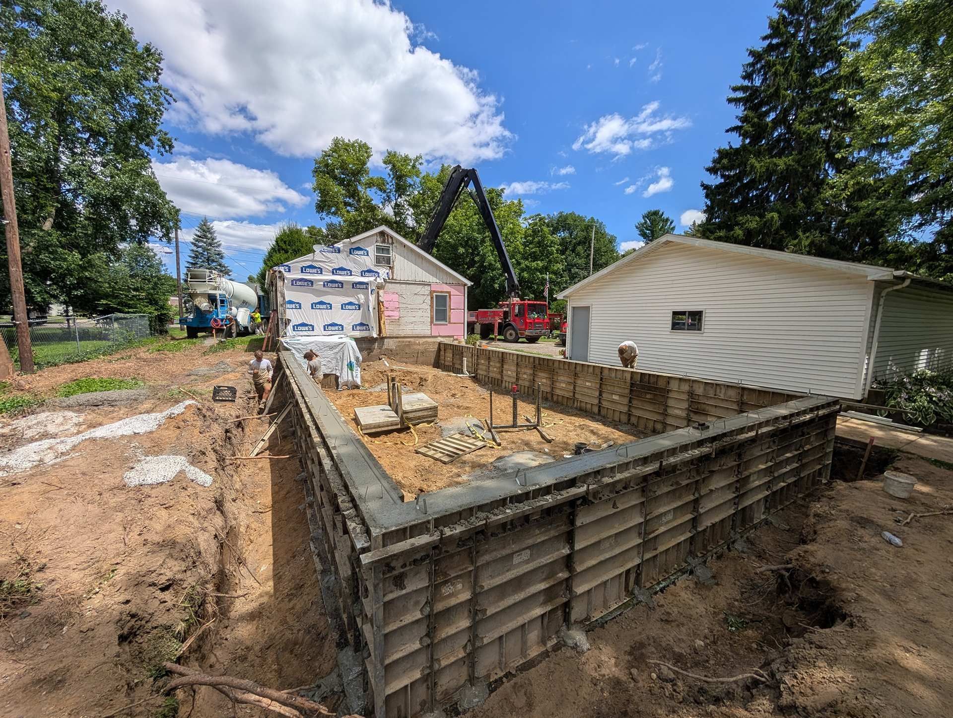 Construction site with a concrete foundation framed by metal forms, adjacent to an existing house and a white garage.