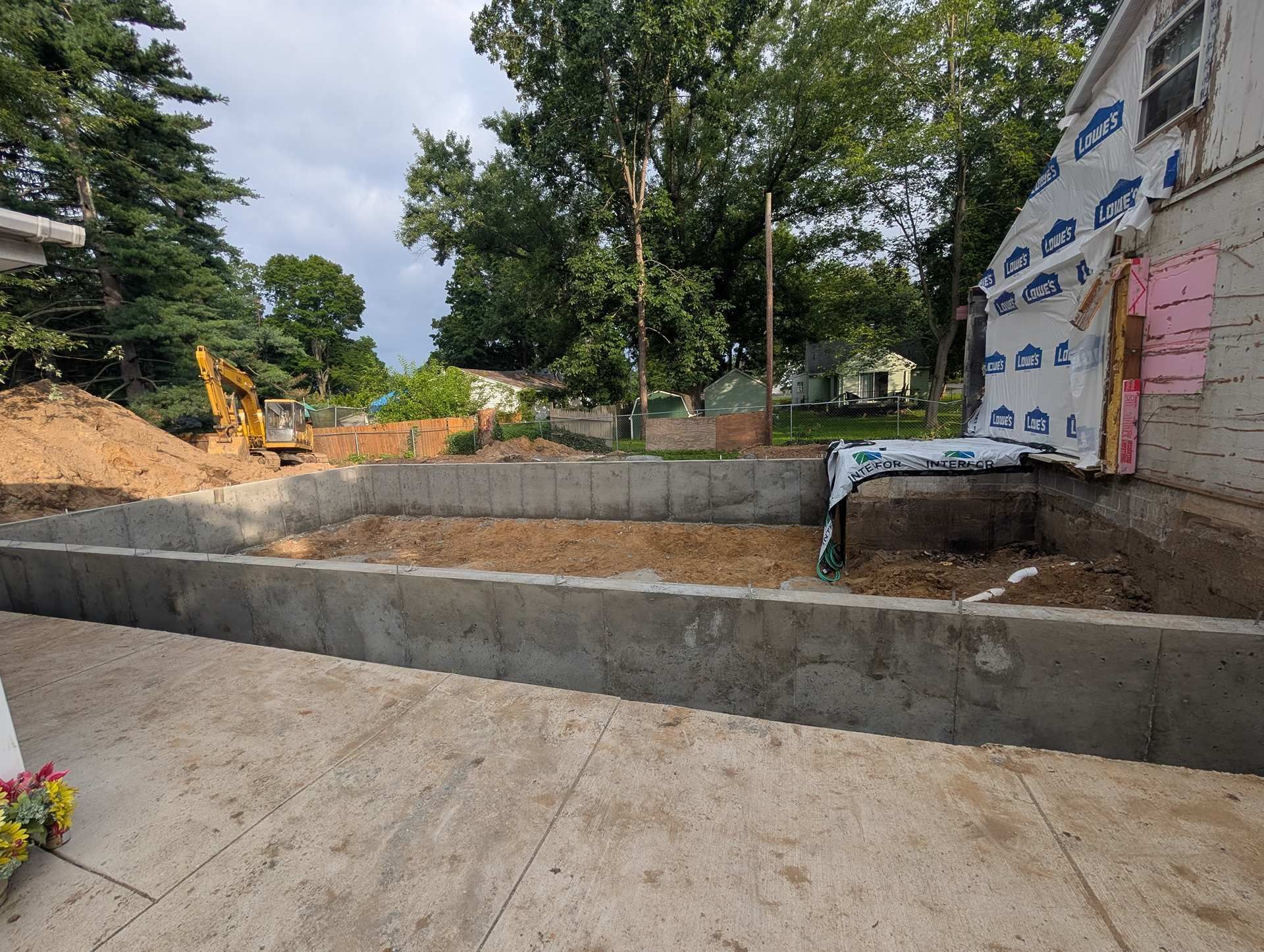 A concrete foundation wall under construction outside a residential home, with a construction site in the background.