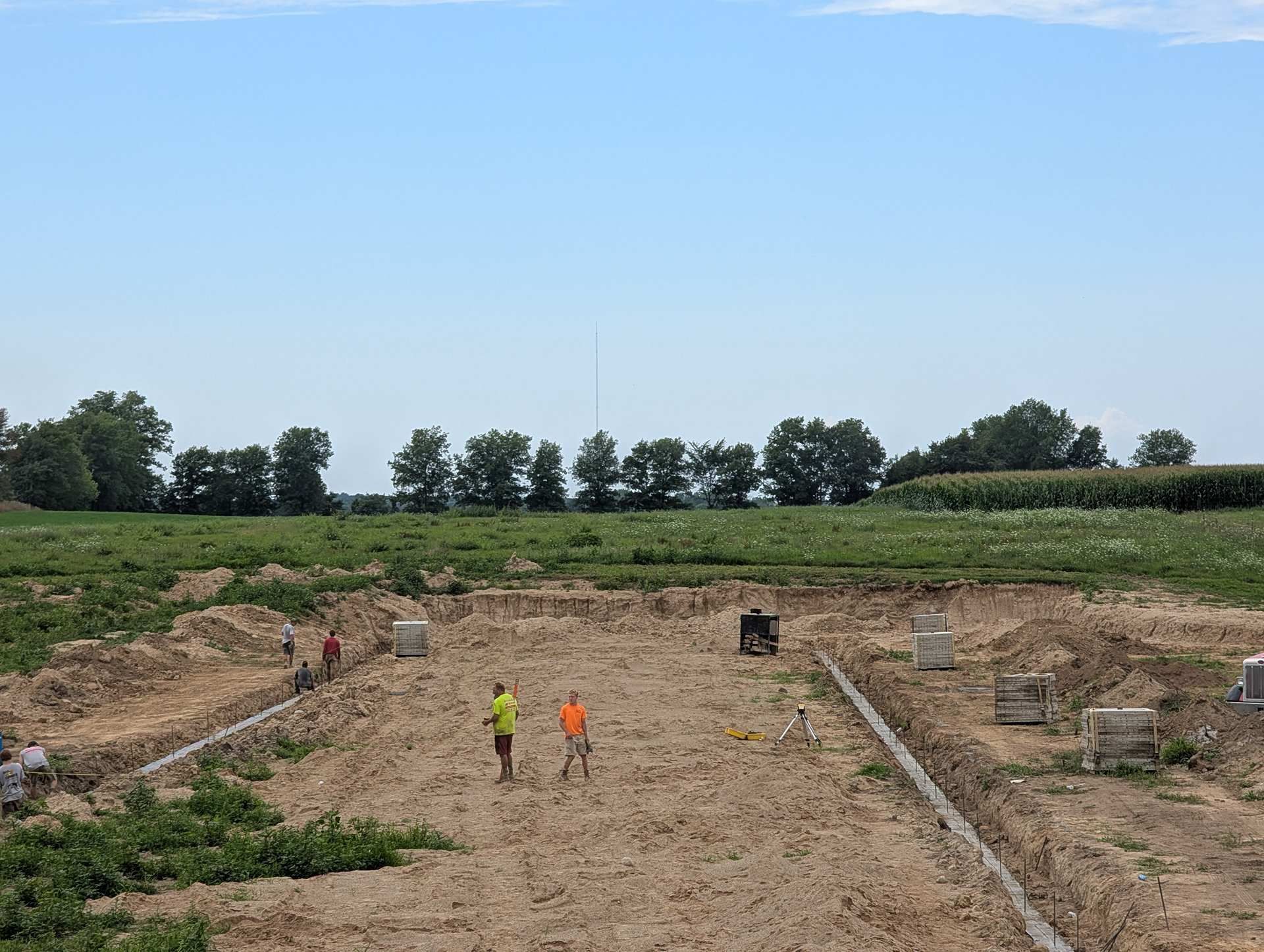 Two people in high-visibility vests walk through a large construction excavation site in a field on a sunny day.