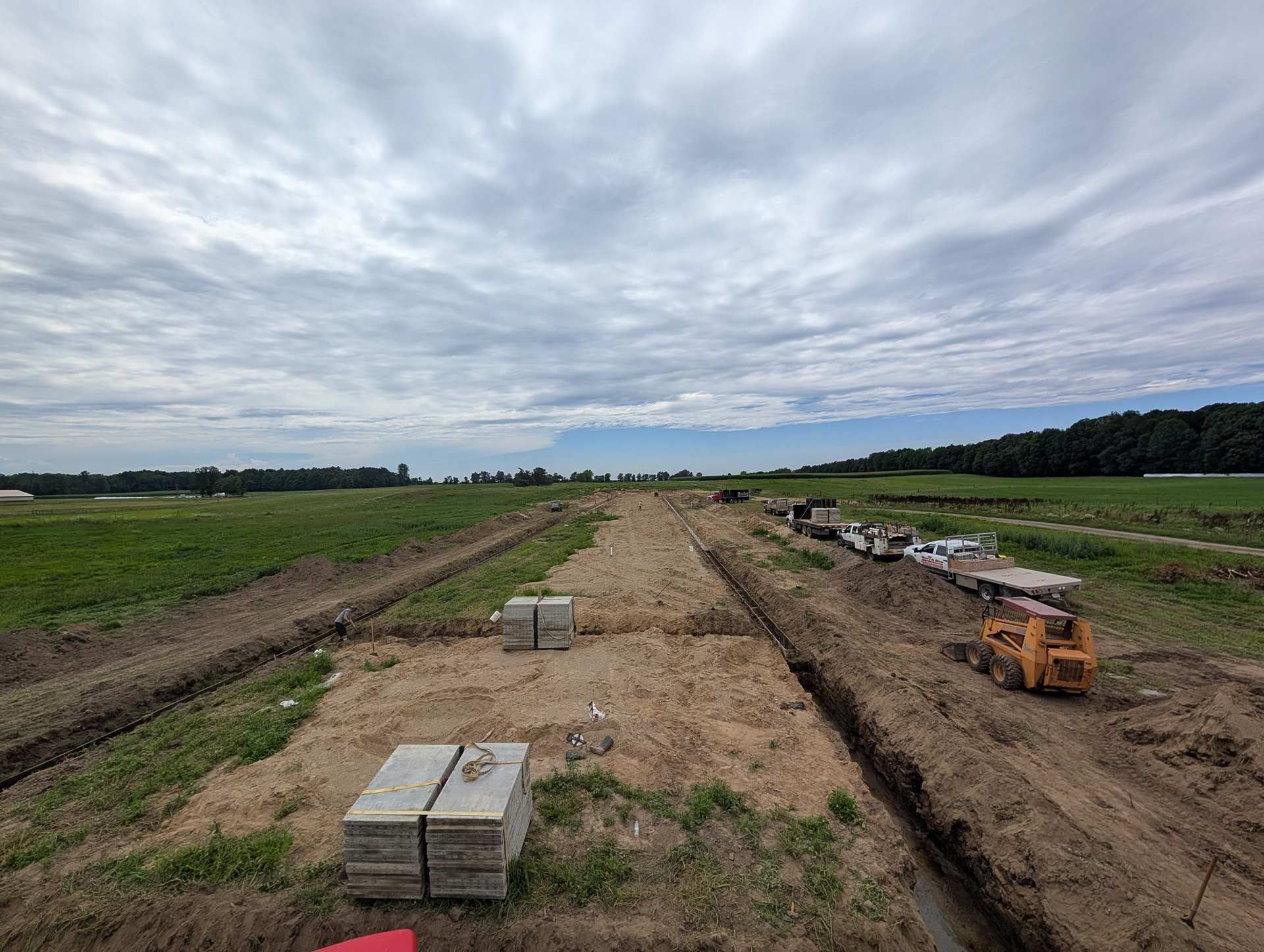 A dirt construction site with parallel trenches, stacks of concrete blocks, and construction equipment under a cloudy sky.