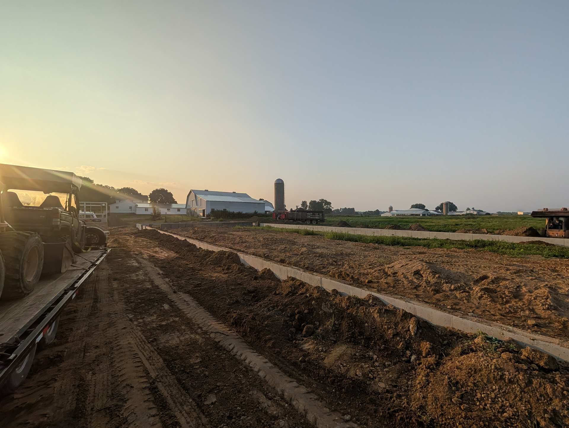 A tractor pulling a trailer across a dirt field at sunset, with farm buildings and a silo in the background.