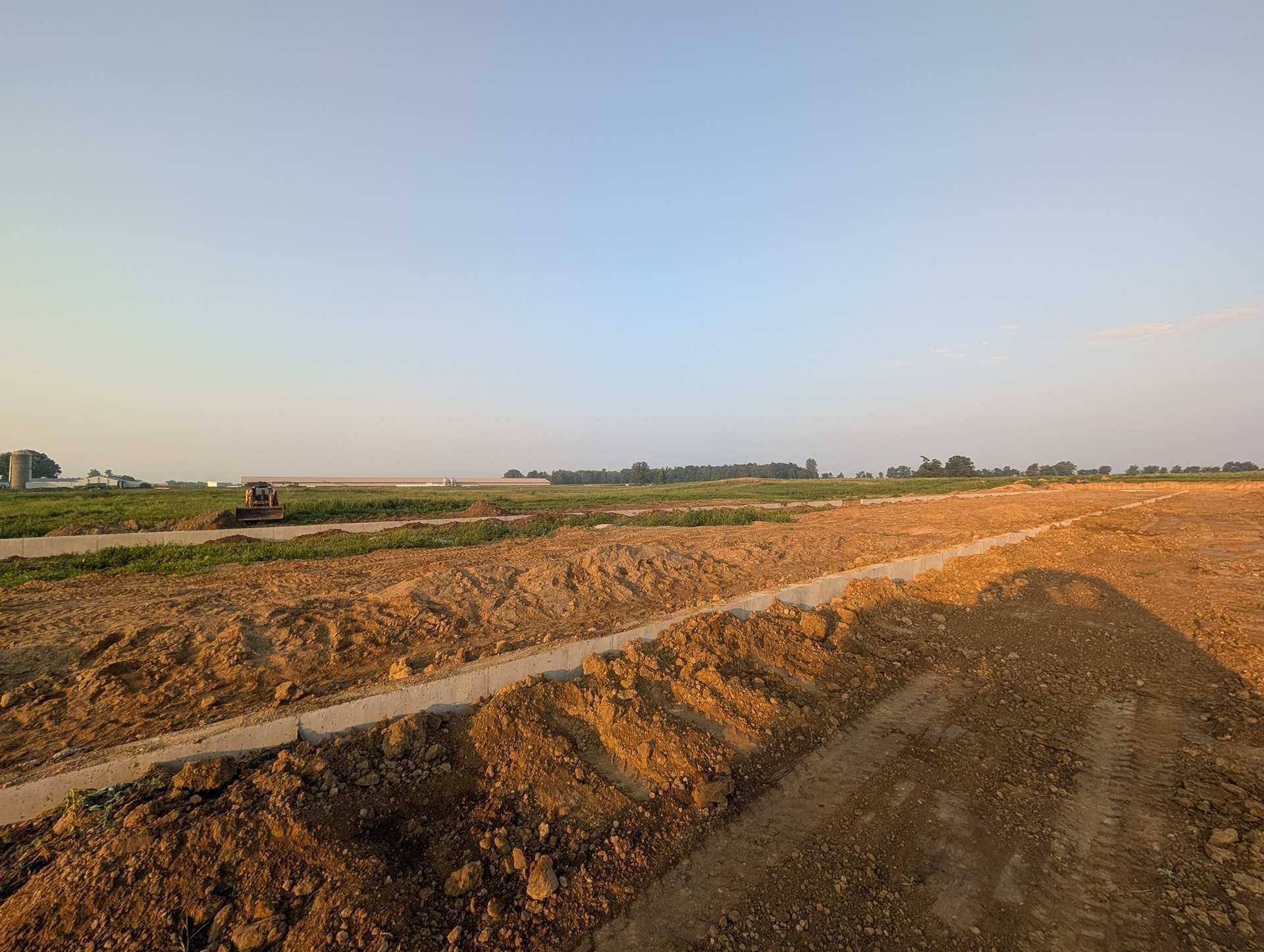 A long, earthen trench stretches through a flat, plowed field under a clear, pale blue sky at sunset.
