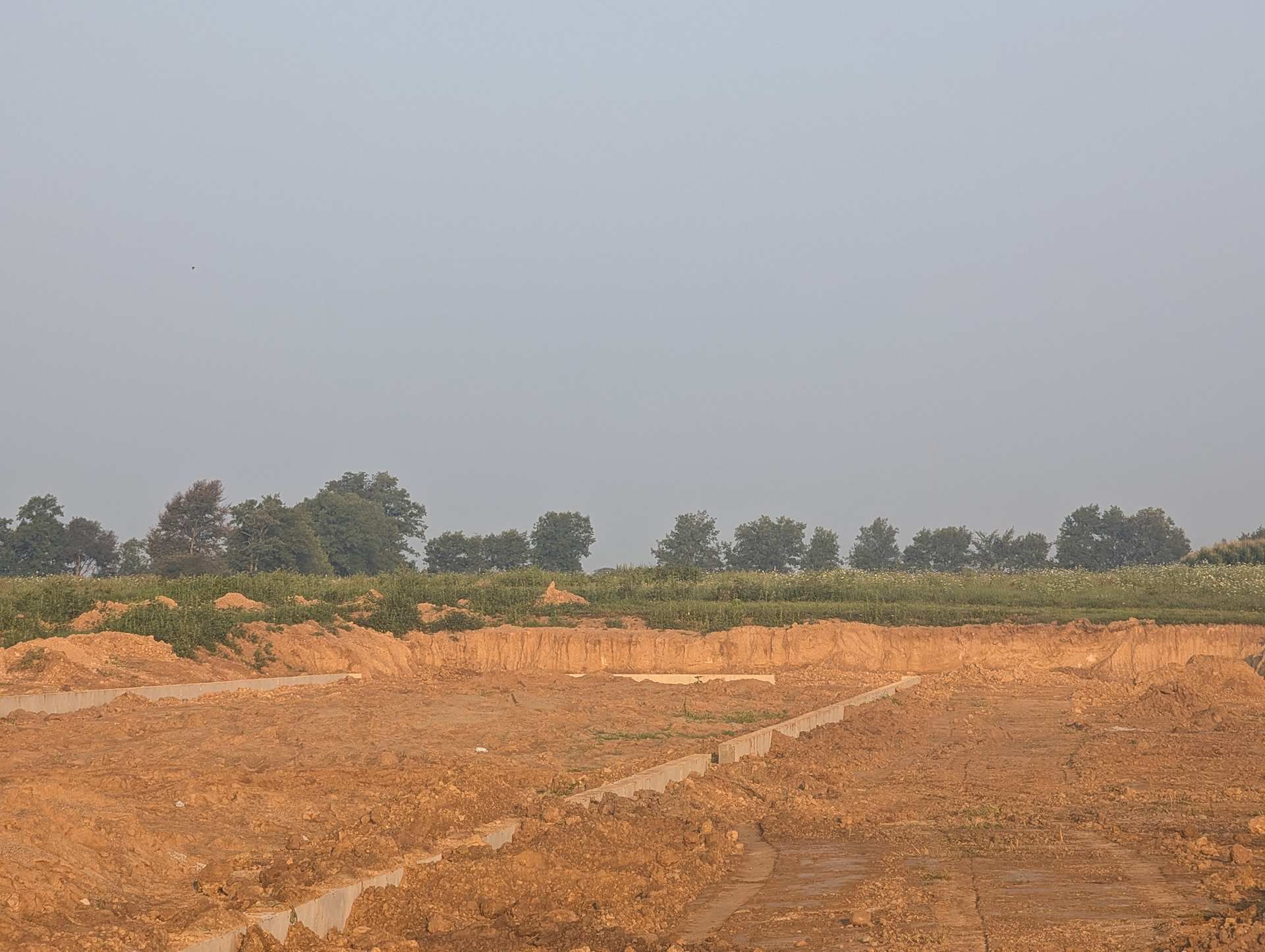 A flat, cleared construction site with reddish-brown soil, bordered by a line of green trees against a hazy, pale sky.