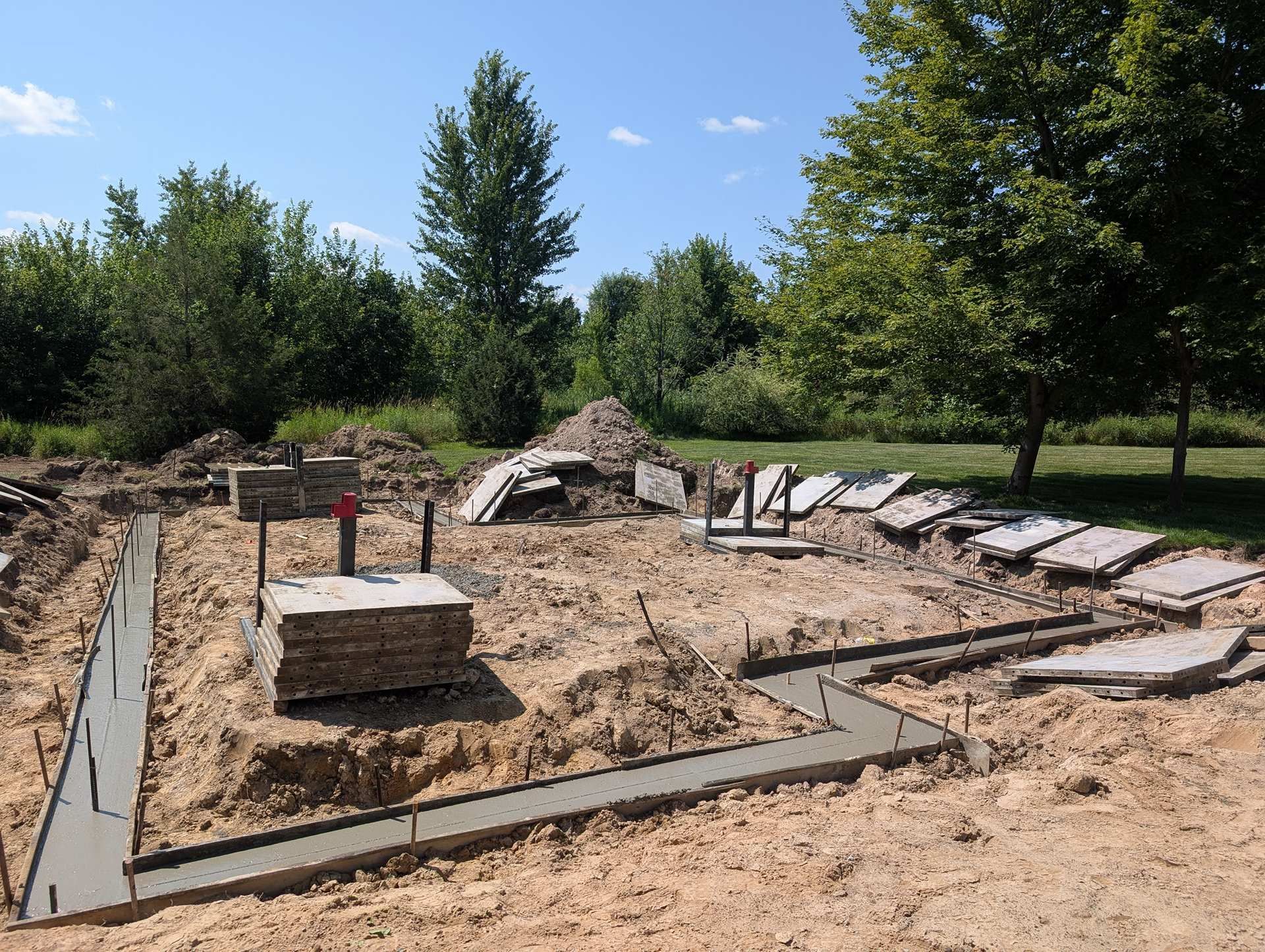 Concrete foundation footings for a building under construction, set in a sunny, grassy field with trees in the background.