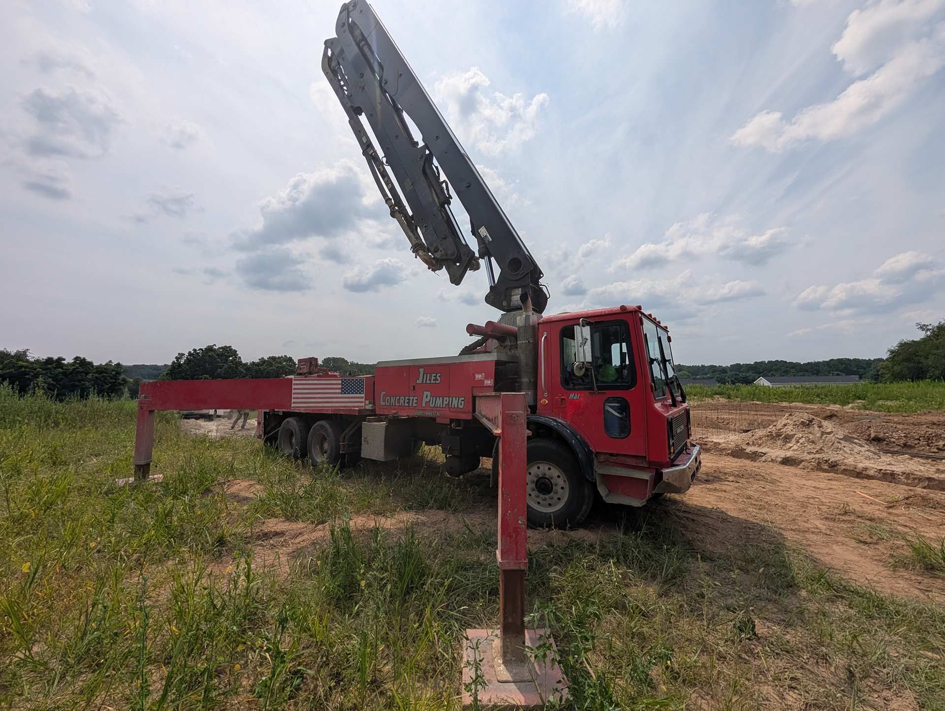 A red concrete pump truck with its boom arm extended, parked on a dirt lot with stabilizer legs deployed.