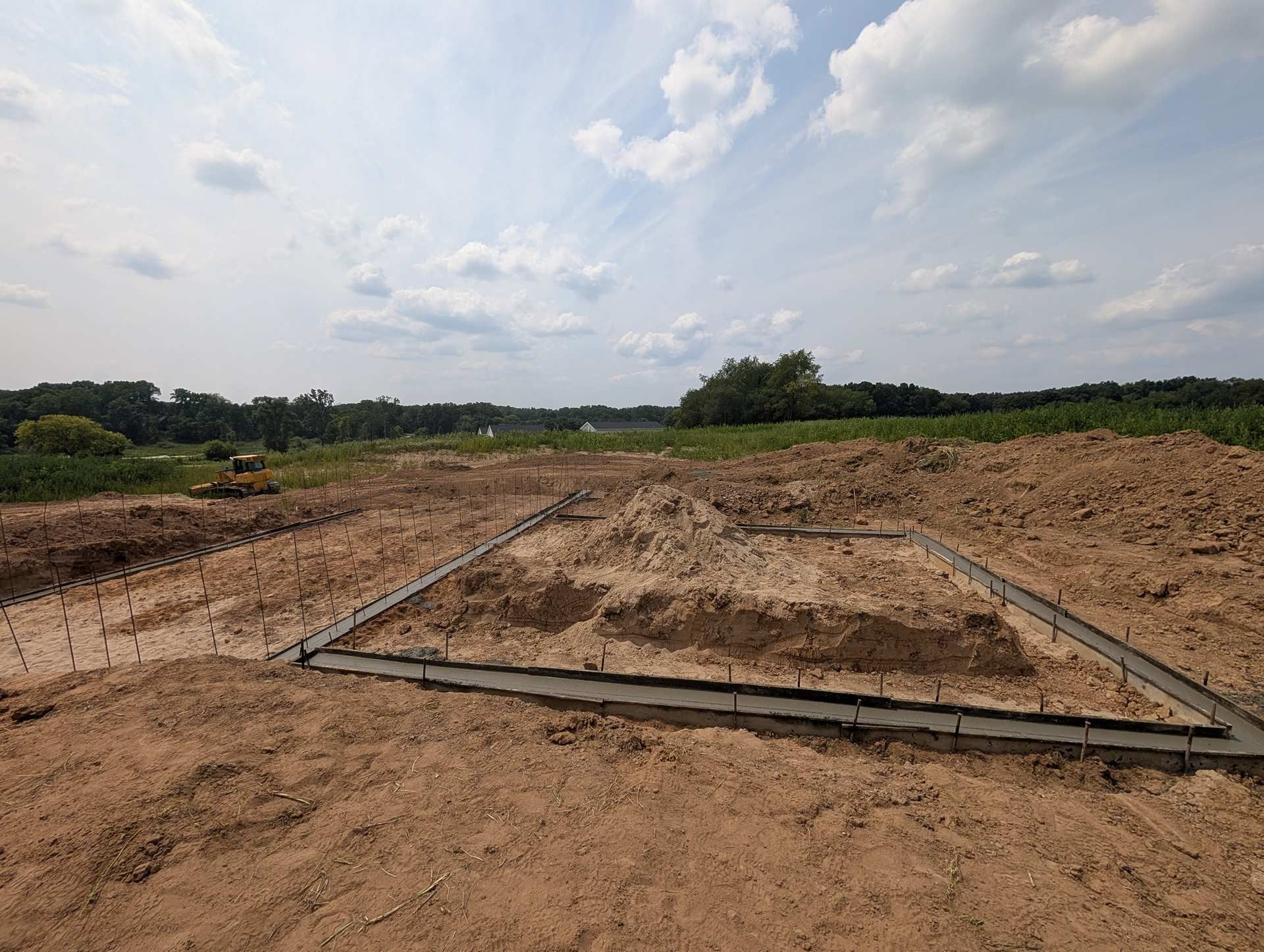 A concrete foundation for a building under construction on a dirt lot, set against a background of trees and a blue sky.