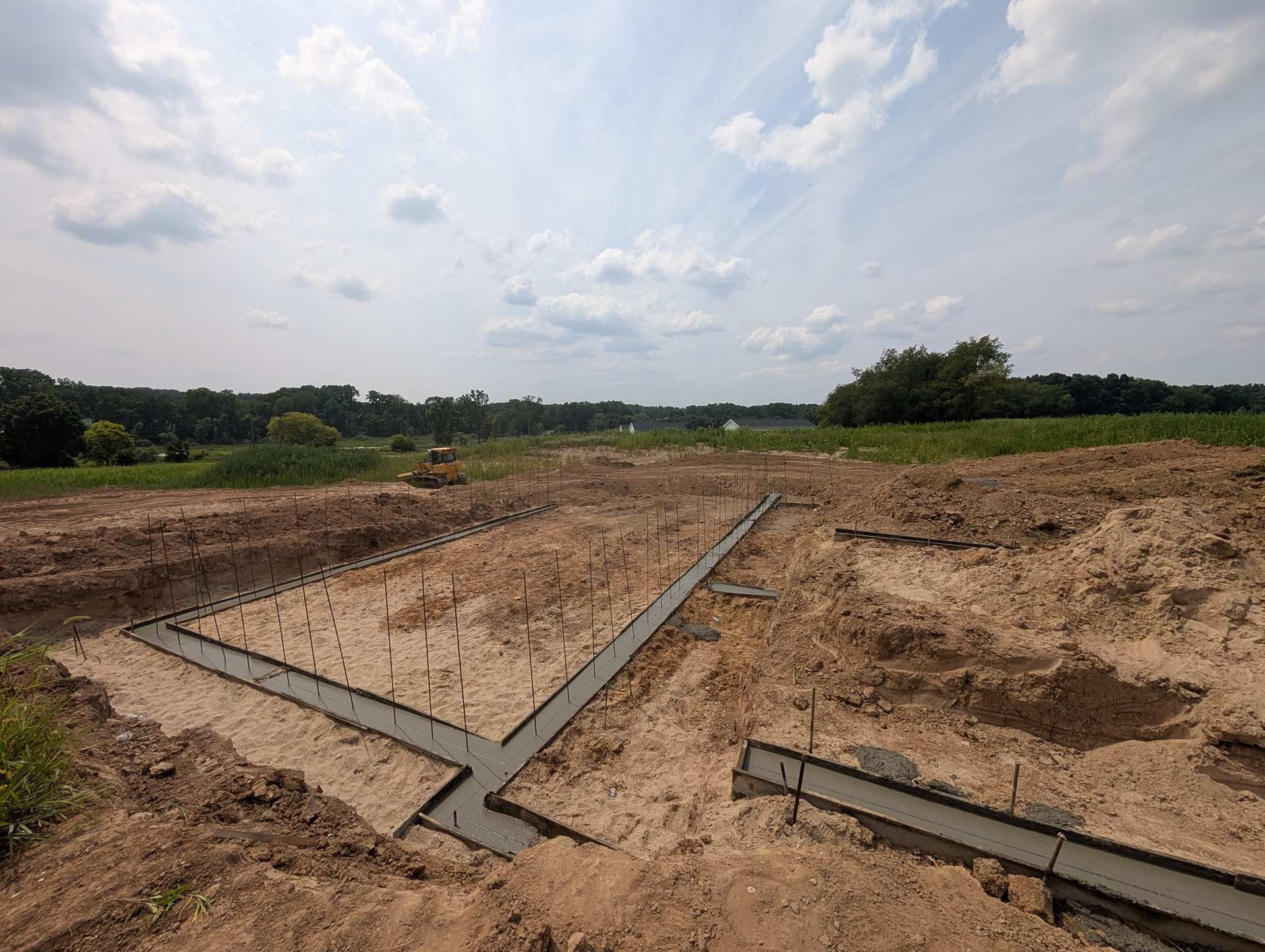 Concrete foundation footings for a building under construction, set in a dirt excavation site under a blue, cloudy sky.