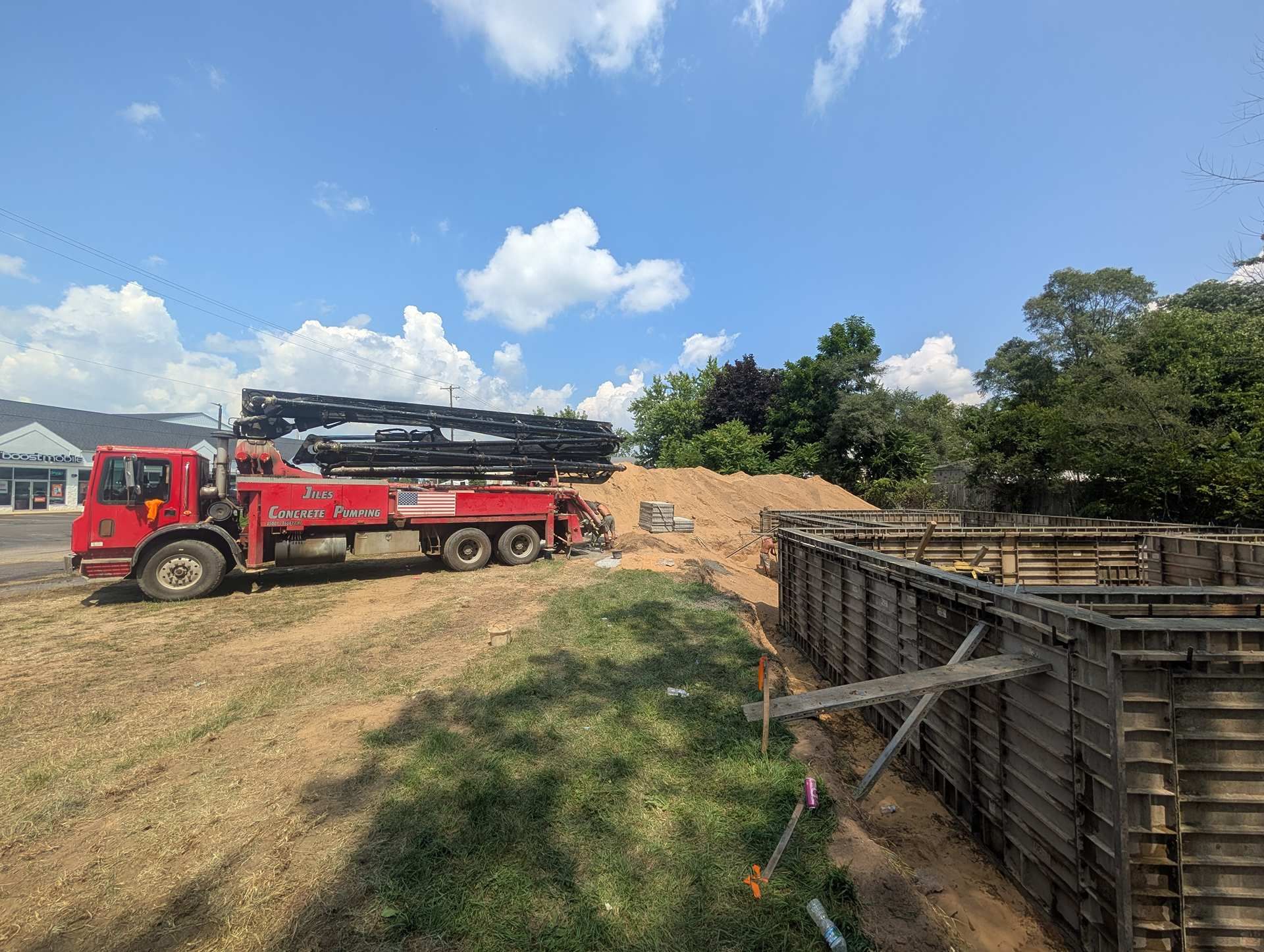 A bright red concrete pump truck parked at a construction site next to wood-framed foundation walls under a blue sky.